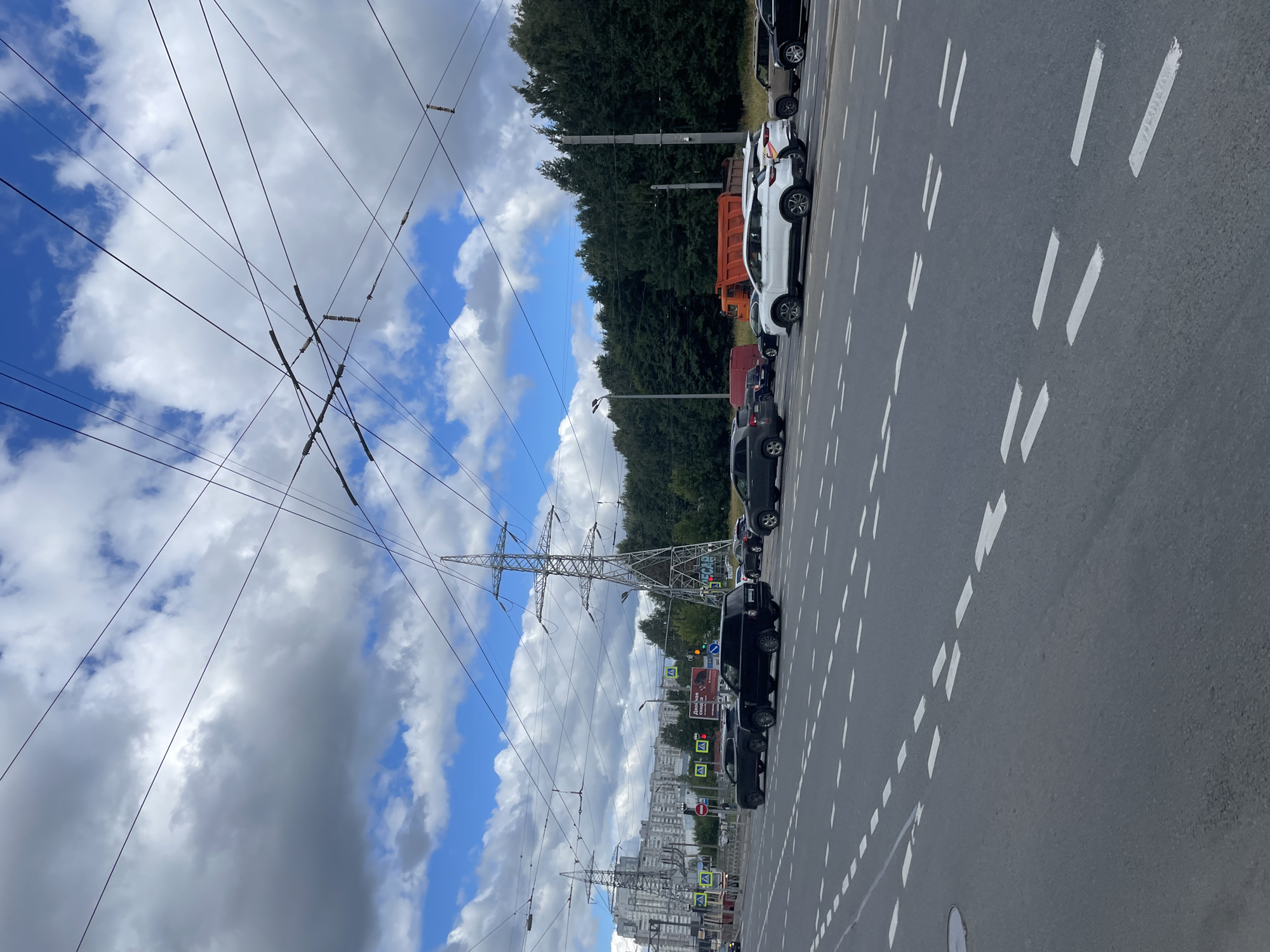 The image depicts a multi-lane asphalt road in Saint Petersburg, Russia, under a cloudy sky with patches of blue. Overhead, a complex network of power lines crisscrosses the frame, supported by tall metal pylons. In the background, dense green trees line the horizon, and a few multi-story buildings are visible on the left. The road is populated with several vehicles, including black SUVs, a white SUV, a red dump truck, and other cars. The road surface is marked with white dashed lines indicating lanes. Traffic signals and streetlights are also present. The scene appears to be a typical urban environment during daylight hours.