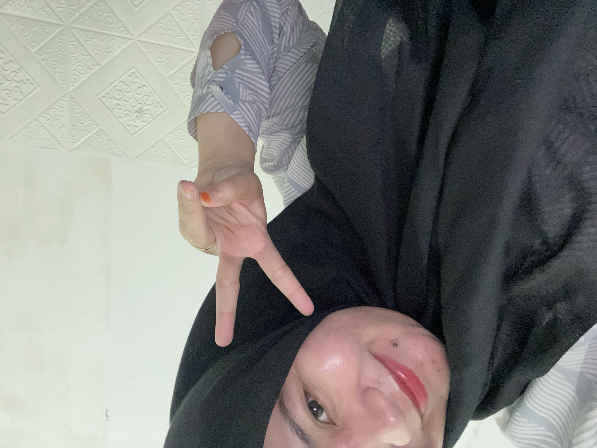 The image shows a close-up selfie of a person wearing a black hijab and a blue and white patterned shirt. The person is smiling and making a peace sign with their right hand. The background appears to be an indoor wall with a textured pattern. The lighting suggests an indoor setting, possibly a home. The specific location in Banjarbaru, Indonesia, cannot be determined from the visual information provided.
