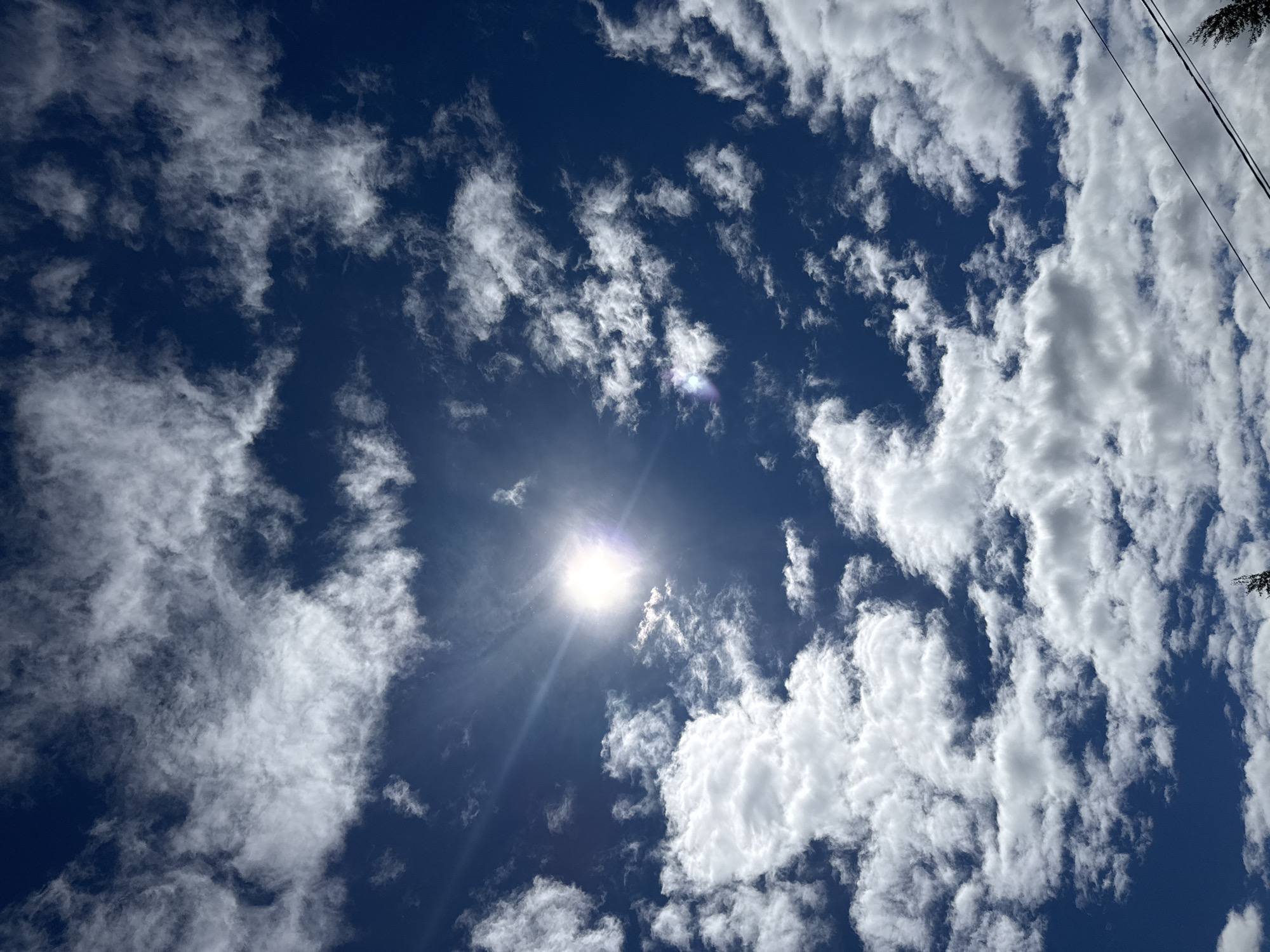 The image shows a bright, clear blue sky with scattered white clouds. The sun is visible, shining brightly in the center of the frame, with sun rays radiating outwards. There are power lines in the upper right corner. The picture was taken outdoors, during the day. It is impossible to confirm the location as Shevlyakovo, Russia from the given image.