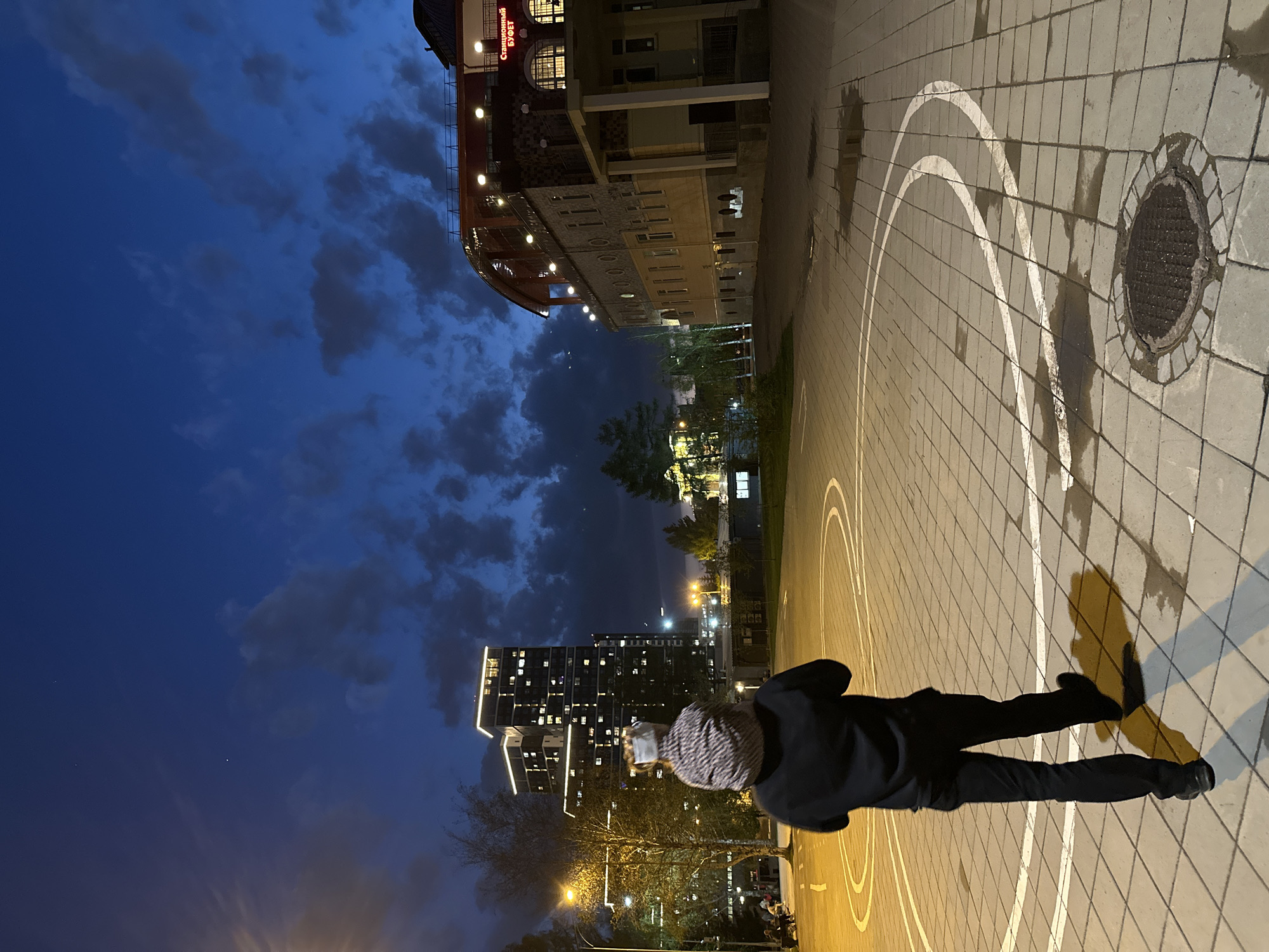 A nighttime shot, rotated 90 degrees clockwise, depicts a person in dark clothing and a patterned covering on their head, walking on a paved area marked with white lines, possibly a basketball court. The person casts a long shadow. The pavement is made of square tiles. A round manhole cover is visible on the right. In the background, buildings are illuminated, including a structure with a curved, lit façade and the inscription "СТАРИННЫЙ БУФЕТ" (Old Buffet) and a tall, modern building with numerous lit windows. Trees and streetlights are scattered in the background. The sky is dark blue with clouds. The location is likely Yekaterinburg, Russia, based on the Cyrillic text on the building.