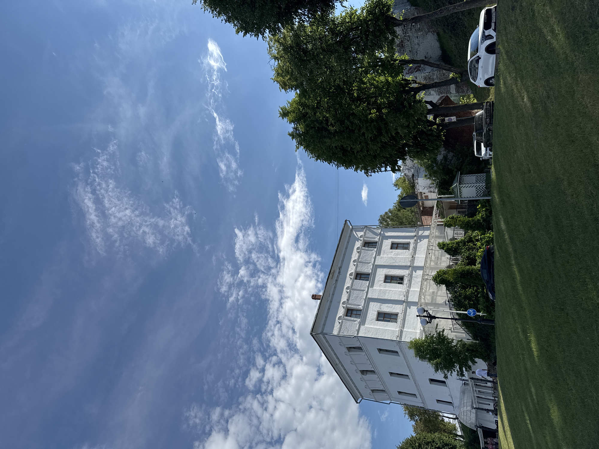This image captures a view of a white, multi-story building with numerous windows, situated on a grassy slope in the city of Zavolzhye, Russia. The building appears to be constructed of brick with a white facade and has a visible chimney on its roof. In front of the building, there is a green lawn, a lamppost, and some foliage. Several cars are parked on a paved area next to the building. The sky above is blue with scattered white clouds. To the right of the building, more trees and a brick wall are visible. A few people are present near the entrance of the building.