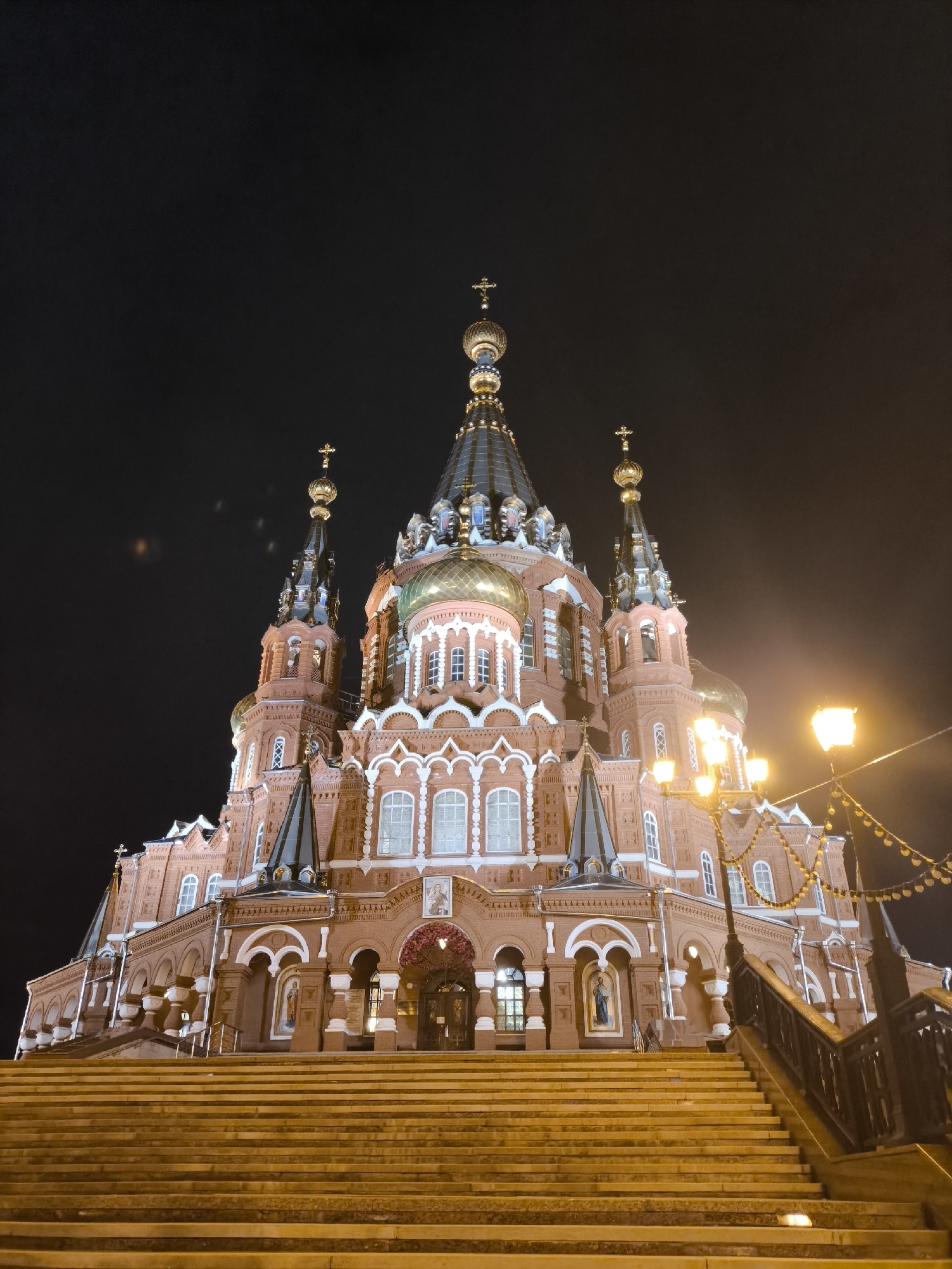 The image depicts Michael's Cathedral (Mikhailovsky Cathedral) in Izhevsk, Russia, at night. The cathedral, constructed primarily from reddish-brown brick with contrasting white decorative architectural elements, dominates the frame, viewed from a low angle. It features multiple golden onion domes, each topped with an Orthodox cross. A large central golden dome is prominent, flanked by several smaller, slender spires also capped with golden elements and crosses. The building exhibits intricate detailing, including arched windows with white frames, decorative cornices, and engaged columns.
A wide, multi-tiered staircase, illuminated with a warm yellow light, leads upwards towards the main entrance of the cathedral. On the right side of the staircase, a black metal railing with decorative elements is visible, ascending alongside the steps. A tall lamppost with multiple glowing yellow lights stands on the right, partially obscured by the cathedral structure, casting a strong light on its immediate surroundings. Decorative string lights or garlands are strung between the lamppost and an unseen attachment point further right. Religious iconography, including painted figures in niches, is visible on the lower facades of the cathedral near the entrance. The sky is dark and appears to be cloudy or hazy, with some minor lens flare visible in the upper left quadrant. No people are present in the scene.