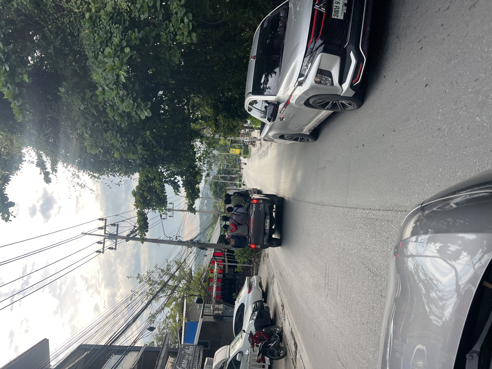 The image shows a street scene in บ้านบางรักษ์, Thailand. A black pickup truck with people riding in the bed is traveling down the road. Parked vehicles and a motorcycle line the side of the street. Overhead power lines run along the left side of the road. A silver pickup truck is visible on the right side of the frame and another pickup truck is visible on the top right of the frame. Trees are visible along the side of the street. Buildings can be seen along the left side of the street.