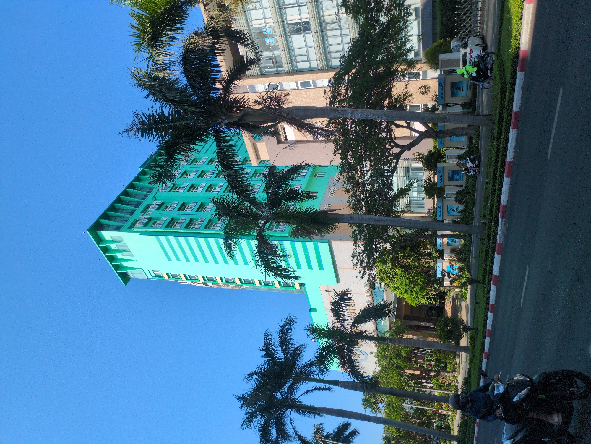A view of the VietBank and CenDeluxe Hotel in Tuy Hòa, Vietnam. The turquoise hotel is visible on the left. The tan VietBank is visible behind a row of palm trees and greenery. A motorcycle is on the right side of the street. A man is riding a motorcycle on the bottom right.
