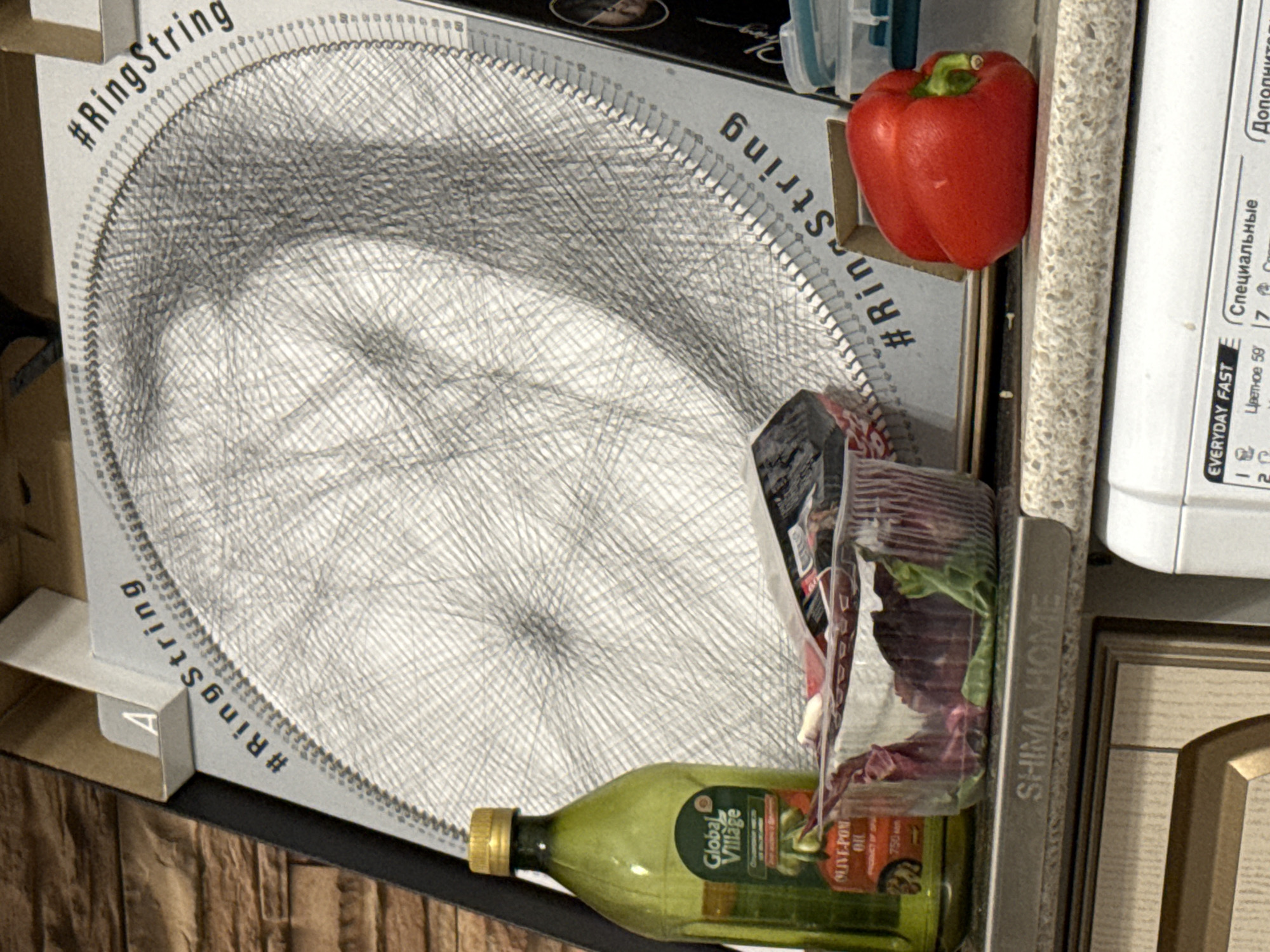 The image shows a cluttered kitchen scene. Dominating the view is a round art piece with a dense web of strings, identified by the hashtag "#RingString" printed around the edge of the frame. Next to it is a red bell pepper, and below are a bottle of Global Village Olive-Pon Oil and a package of greens. To the right, a white appliance with text in Russian ("Специальные", "Дополнительные") is visible. The countertop has a speckled pattern. A metal bar labeled "SHIMA HOME" is placed on the front of the cabinets. The kitchen cabinets have a light wood finish. This image does not contain information to provide the location in Izhevsk, Russia.