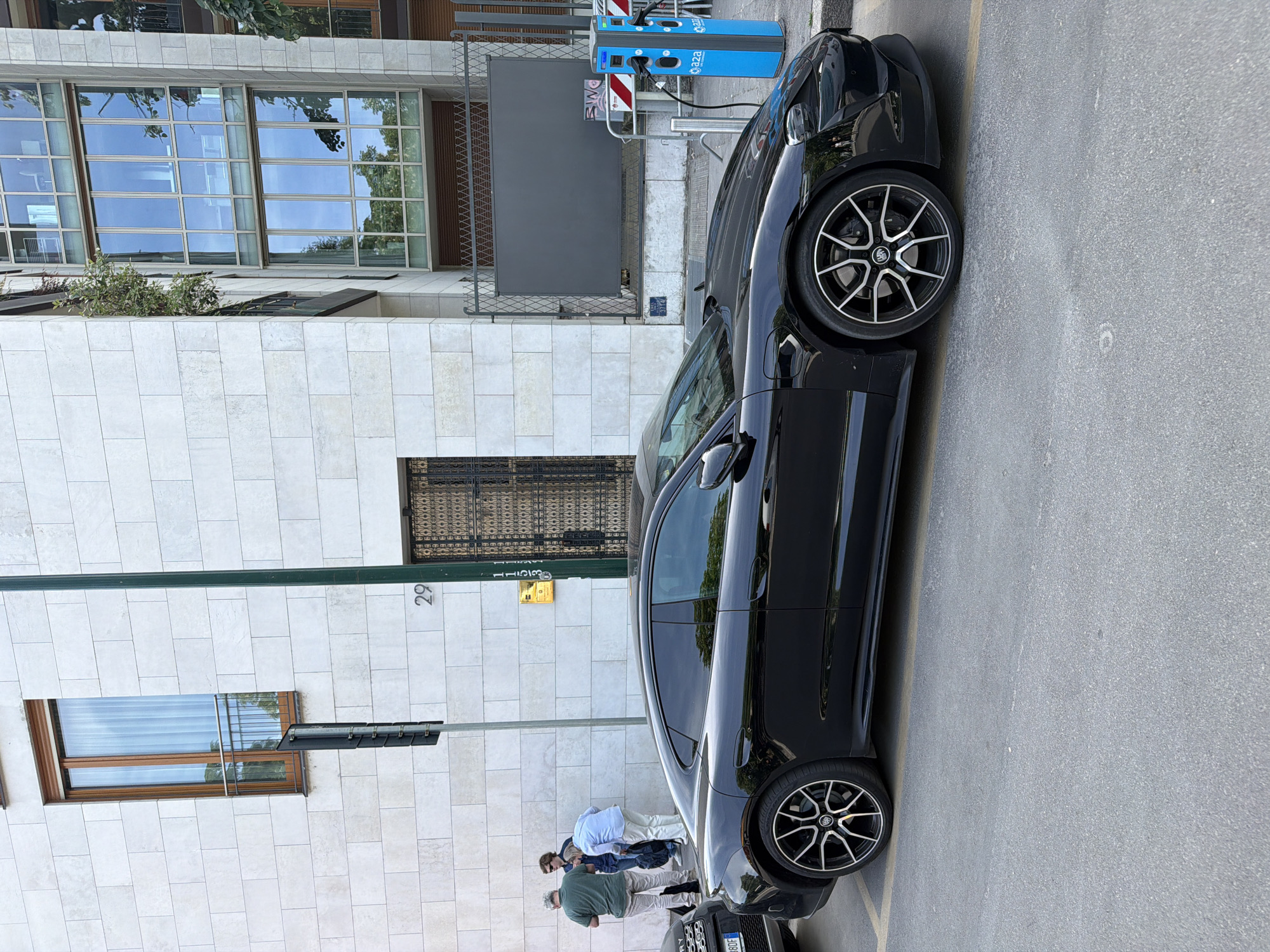 The image captures a scene on a street in Milan, Italy. A black car is parked next to a building with light-colored stone blocks and windows. There are three people standing on the sidewalk near the rear of the car. An electric vehicle charging station is visible next to the car. The image is slightly rotated.