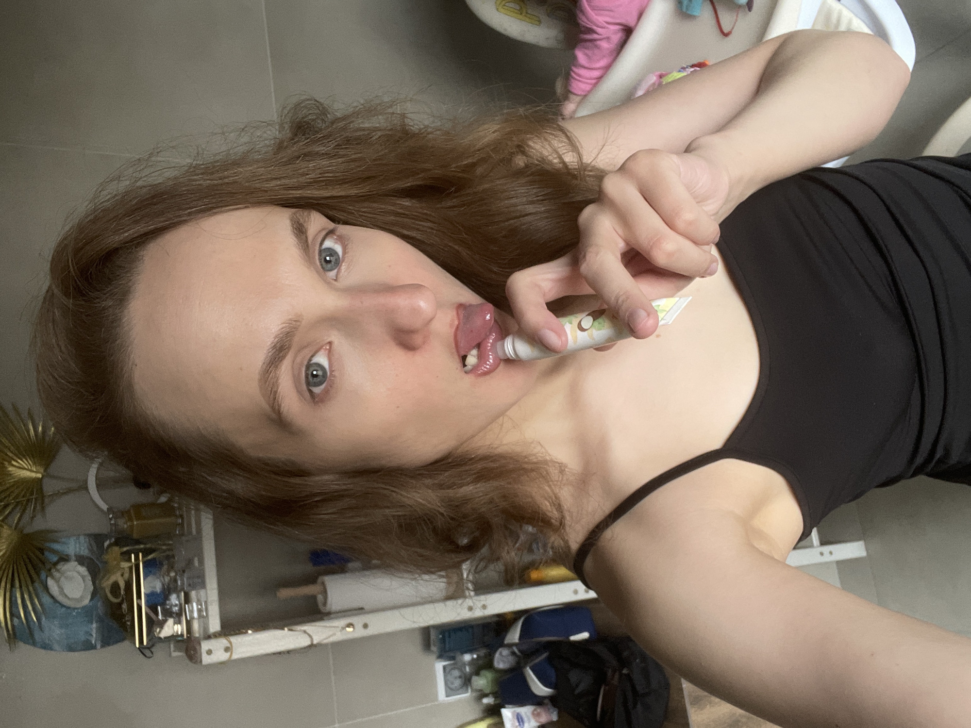The media file presents a rotated, close-up, selfie-style photograph taken indoors in Подшивалово, Russia. The central subject is a young individual, likely female, with shoulder-length wavy brown hair and piercing blue eyes that look directly at the viewer. She is dressed in a plain black tank top or camisole, revealing her fair skin.
The person is engaged in an activity involving a small, white tube with a distinctive green and white logo, which appears to feature a coconut or a leaf design. She holds the tube up to her mouth, with her tongue slightly extended, making contact with the tube's opening. This action suggests she is either applying a product like lip gloss or lip balm, or perhaps consuming a liquid snack from the tube. Her expression is a mix of concentration and playful candor, highlighted by her wide, direct gaze.
The background elements indicate a lived-in, domestic setting. To the left, a cluttered shelf or surface is visible, holding a unique gold-colored, fan-shaped decorative item, along with various bottles and small, indistinct objects. A roll of white material, possibly paper towels or cling film, is also present on this shelf. In the upper right corner of the image, there are faint visual cues suggesting the presence of a child, including a pink-sleeved arm and a partial view of what might be a crib or playpen structure. The surrounding walls or floor are a neutral grey color. While a logo is visible on the tube, specific readable text is not discernible. The overall atmosphere captured is an intimate and candid moment within a personal home environment.