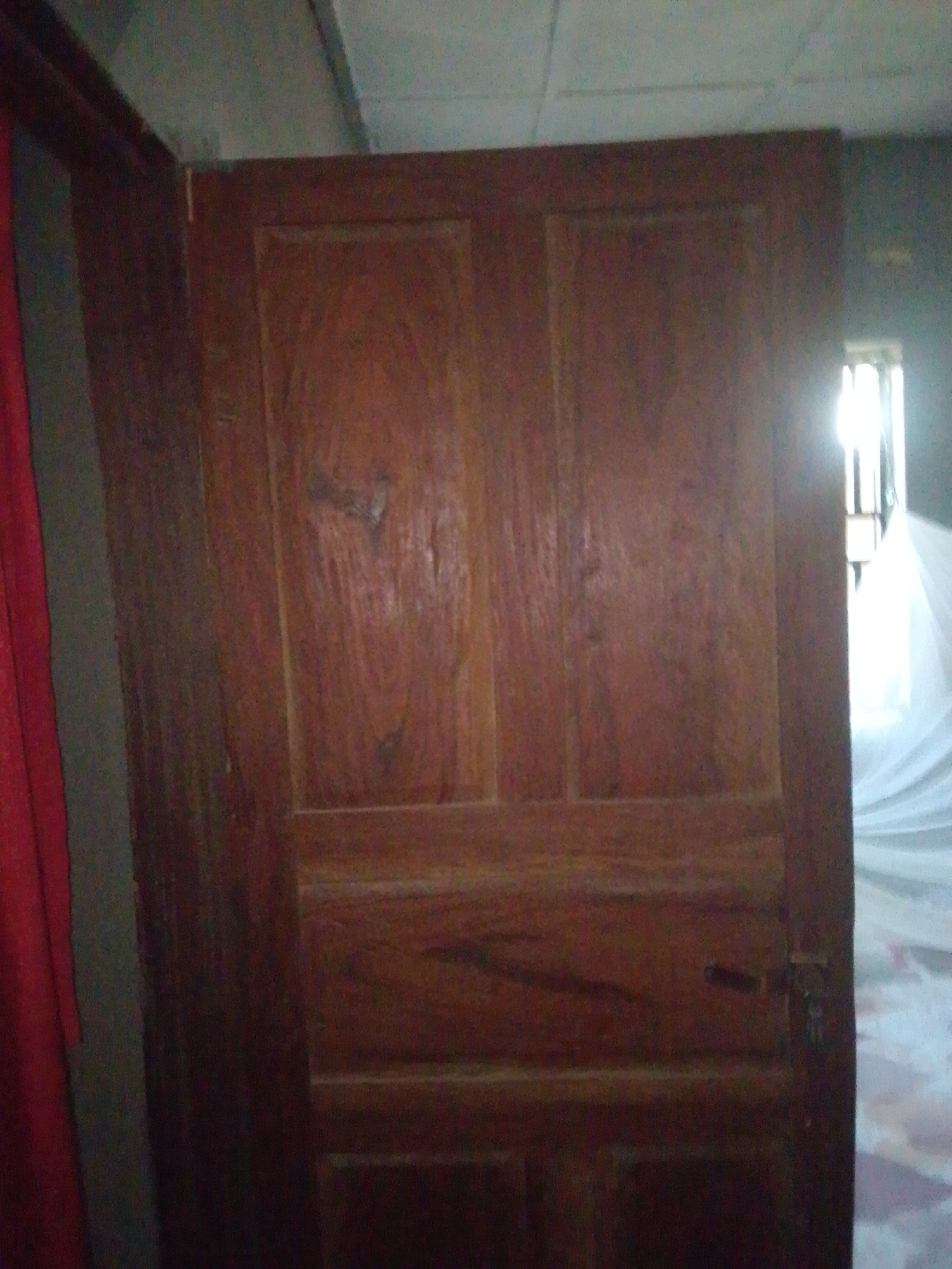 The image shows a wooden door, likely an interior door, with a visible latch and hinge. The door has a paneled design with horizontal and vertical lines creating distinct sections. The wood grain is visible, suggesting it is a natural wood material with a warm, brown tone. To the left of the door, a glimpse of a red curtain or fabric can be seen, partially obscuring a wall or another surface. To the right of the door, a bright light source is visible, possibly a window, with a white, sheer material draped in front of it, creating a diffused light effect. The overall impression is of an indoor setting. The context provided indicates the location is Jalingo, Nigeria. No people are visible in the image. The lighting suggests it could be daytime, as there is a strong light source coming from what appears to be a window. No specific activity or event is depicted, as the image focuses on the door and its immediate surroundings. There is no visible text in the image.