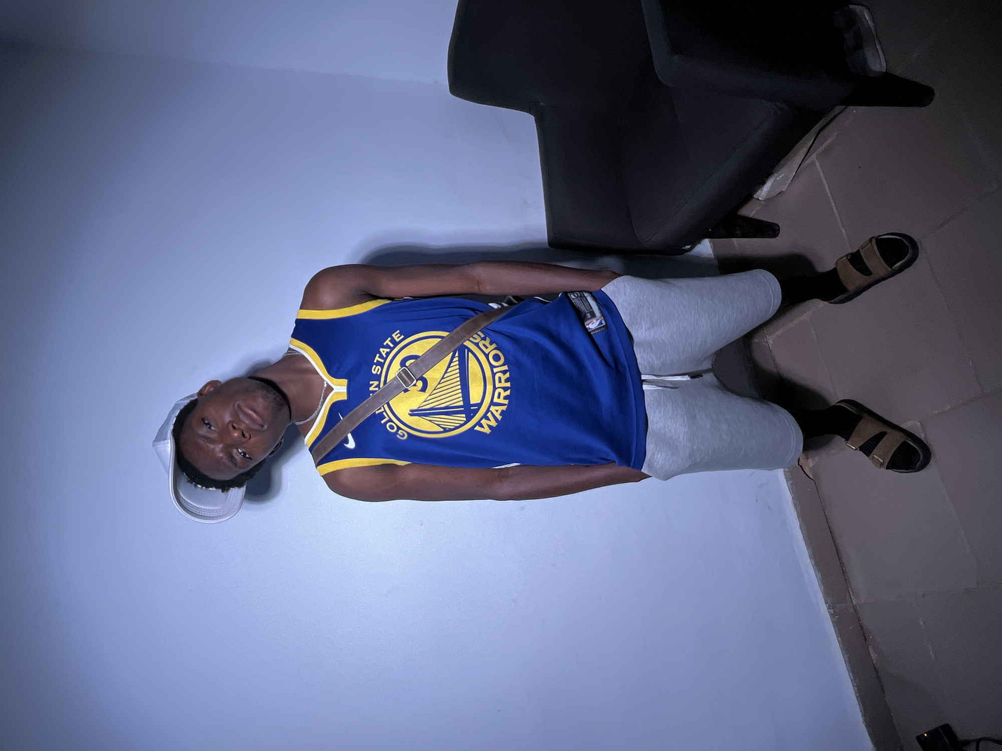 A person, likely male, is captured in a vertical, slightly tilted photograph against a light blue wall. They are wearing a blue and gold Golden State Warriors basketball jersey over a white t-shirt, light gray sweatpants, and black socks with brown slides. A brown strap, possibly from a bag, crosses their chest. They are also wearing a white baseball cap. The individual is looking towards the camera with a neutral expression. In the background, a dark-colored chair is visible, partially obscuring the wall. The floor appears to be tiled in muted tones. The lighting suggests it could be indoors, potentially in the evening or a room with artificial lighting, as there are distinct shadows and a spotlight effect on the wall. No specific time of day or weather conditions can be determined. Visible text on the jersey reads "GOLDEN STATE WARRIORS".