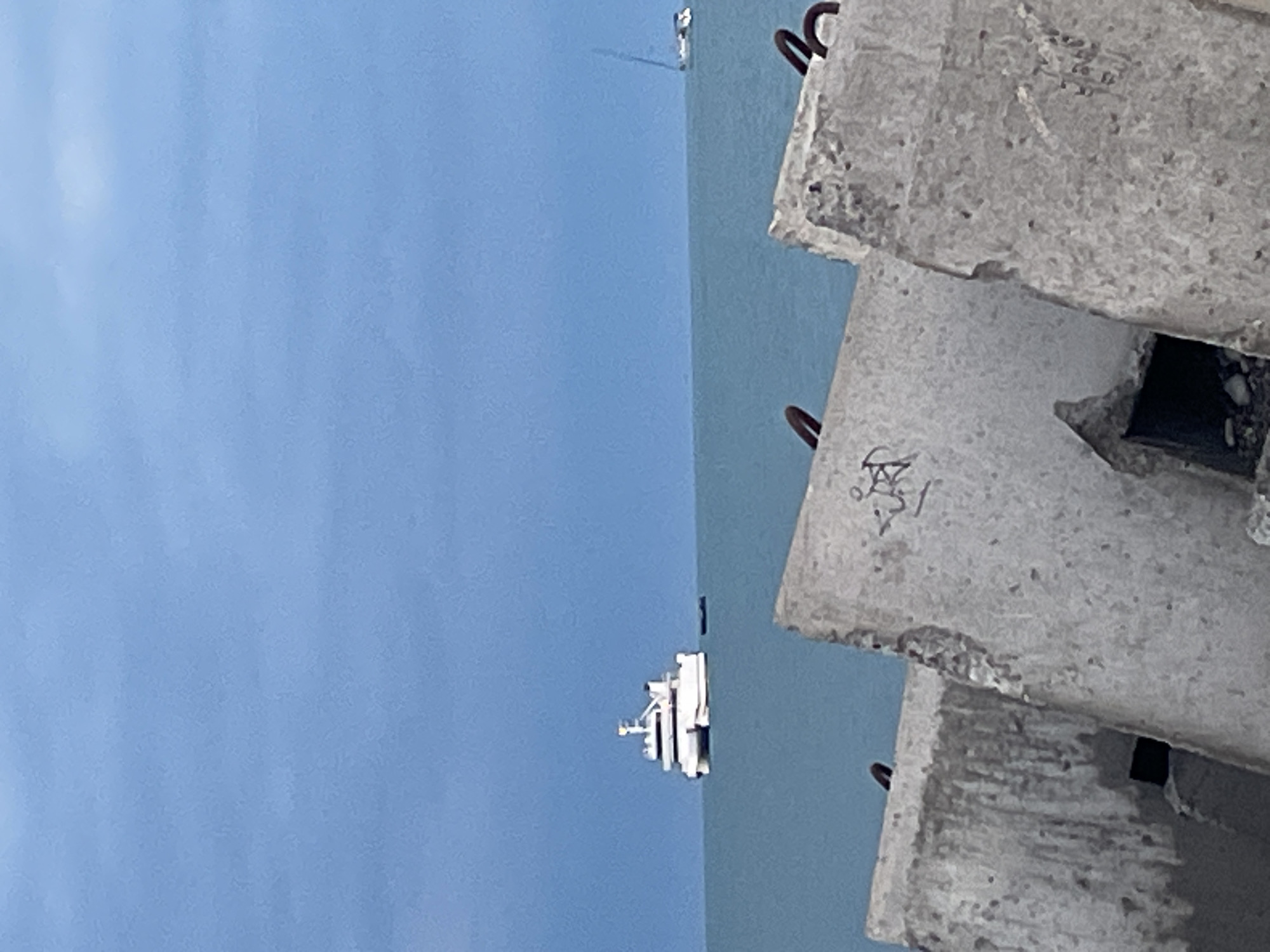 In a wide shot, a calm blue ocean under a hazy, pale blue sky. In the mid-ground, a white, multi-decked catamaran sits on the water. To the right, a white sailboat with a single mast is visible in the distance. In the foreground, large, weathered concrete blocks are arranged, some with rusty metal rings protruding from the top and some showing signs of graffiti. The scene is an outdoor, seaside environment.