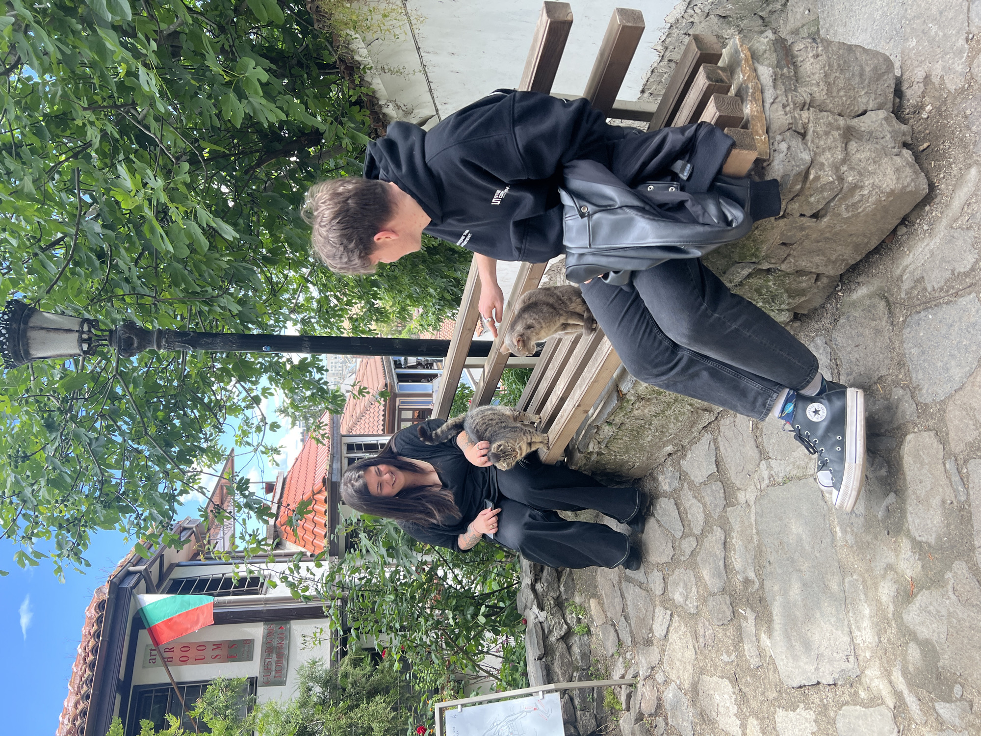 Two people, one male and one female, are interacting with cats while seated on a stone and wood bench in Plovdiv, Bulgaria. The man, wearing a black hoodie and jeans with black Converse sneakers, is seated on the bench, petting a cat on his lap. The woman, wearing a black shirt and pants, is sitting on the ground with a cat in her arms. In the background, a lamppost, lush green trees, a building with a Bulgarian flag, and a stone-paved ground can be seen.