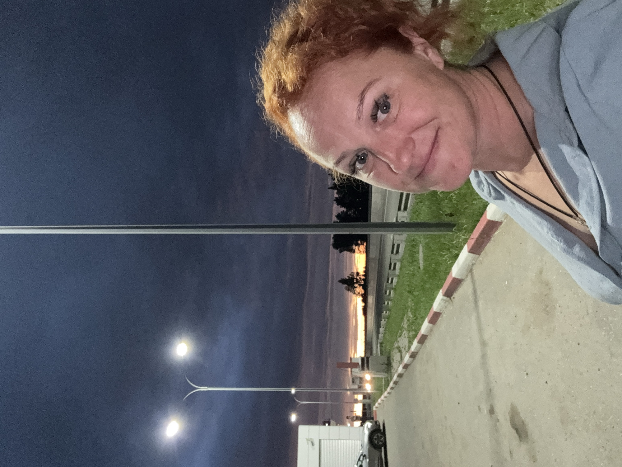 A woman with red curly hair is taking a selfie outdoors at dusk. She is wearing a light blue hooded sweatshirt. Behind her, a street light illuminates a paved area with a white and red striped curb. A car is parked to her left. The sky is dark blue with dramatic clouds, and a faint sunset glow can be seen on the horizon. In the distance, trees and some buildings are visible. The location is likely a roadside, possibly a gas station or rest stop, in the city of Можга, Russia.