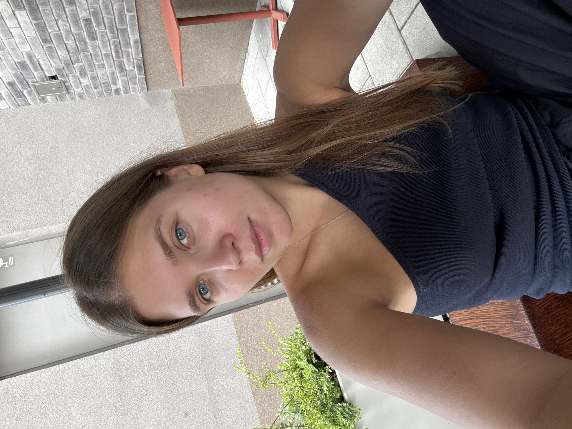 A young woman with long brown hair and blue eyes takes a selfie outdoors. She is wearing a dark blue, ribbed, one-shoulder top. Her skin has a tan appearance with some light freckles. The background shows a textured light-colored wall to her left, a planter with green foliage, and a brick wall to her right. A small, orange table is visible in the background. The setting appears to be an outdoor seating area, possibly a cafe or restaurant, in Belgrade, Serbia.