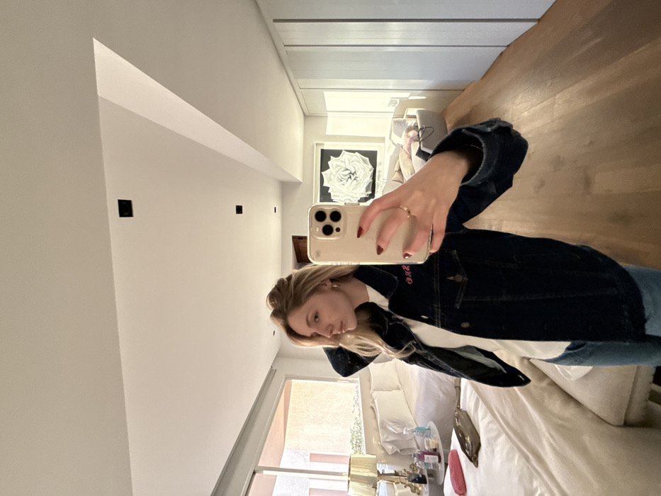 The scene depicts a mirror selfie taken by a fair-skinned individual with long blonde hair, dressed in a white t-shirt and a dark denim jacket featuring pink embroidery (possibly "ONZ" or "2N0") on the left chest. The individual holds a light-colored smartphone with a clear case in their left hand, displaying dark red nail polish and a gold ring on the ring finger, while their head is turned to the left in a profile view. A gold hoop earring is visible on the left ear.
The image is a reflection of a modern, well-lit bedroom or master suite. To the left, a large window or glass door provides natural light and a view of light-colored, multi-story buildings. Adjacent to the window is a large bed with white bedding and multiple pillows. A bedside table holds an ornate gold lamp, several small bottles, and a pink pouch. The room features light wooden flooring and a white ceiling with black recessed light fixtures and a linear lighting element. Along the right side of the room, a wall of white built-in wardrobes or cabinets is visible. Further into the room, a light-colored sofa or sectional is present, with a dark item draped over it, and a large framed artwork featuring a white, rose-like floral design on a dark background hangs on the wall.
The location is within an upscale residential property in Mexico City, Mexico. While a specific colonia or address is not discernible, the contemporary interior design, spacious layout, and exterior view of similar affluent residential architecture are consistent with high-end neighborhoods such as Polanco, Lomas de Chapultepec, or Santa Fe.