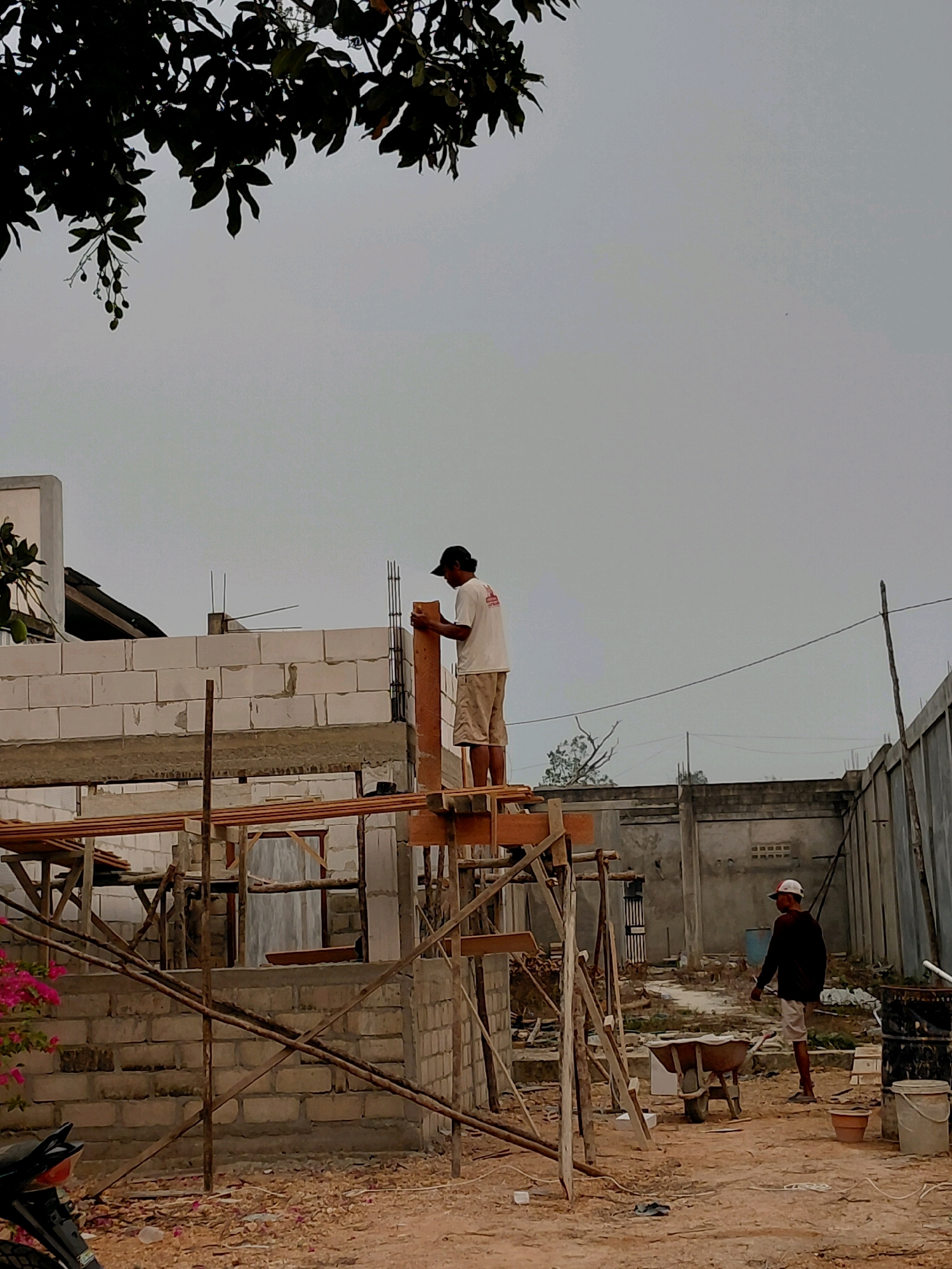 The image depicts a construction site in Persiapan Mulya Raya, Indonesia, under an overcast sky. In the foreground, a partially constructed building is visible, made of white cinder blocks and reinforced with wooden scaffolding. A person wearing a white t-shirt and shorts, with a black cap, is standing on the scaffolding, holding a wooden plank. Further in the background, another person in a dark shirt, white shorts, and a white cap is walking towards the right side of the frame. The ground is covered with dirt and debris. Various construction materials and tools, including a wheelbarrow, buckets, and pipes, are scattered around the site. The surrounding environment appears to be a developing area with other buildings and fences in the distance.