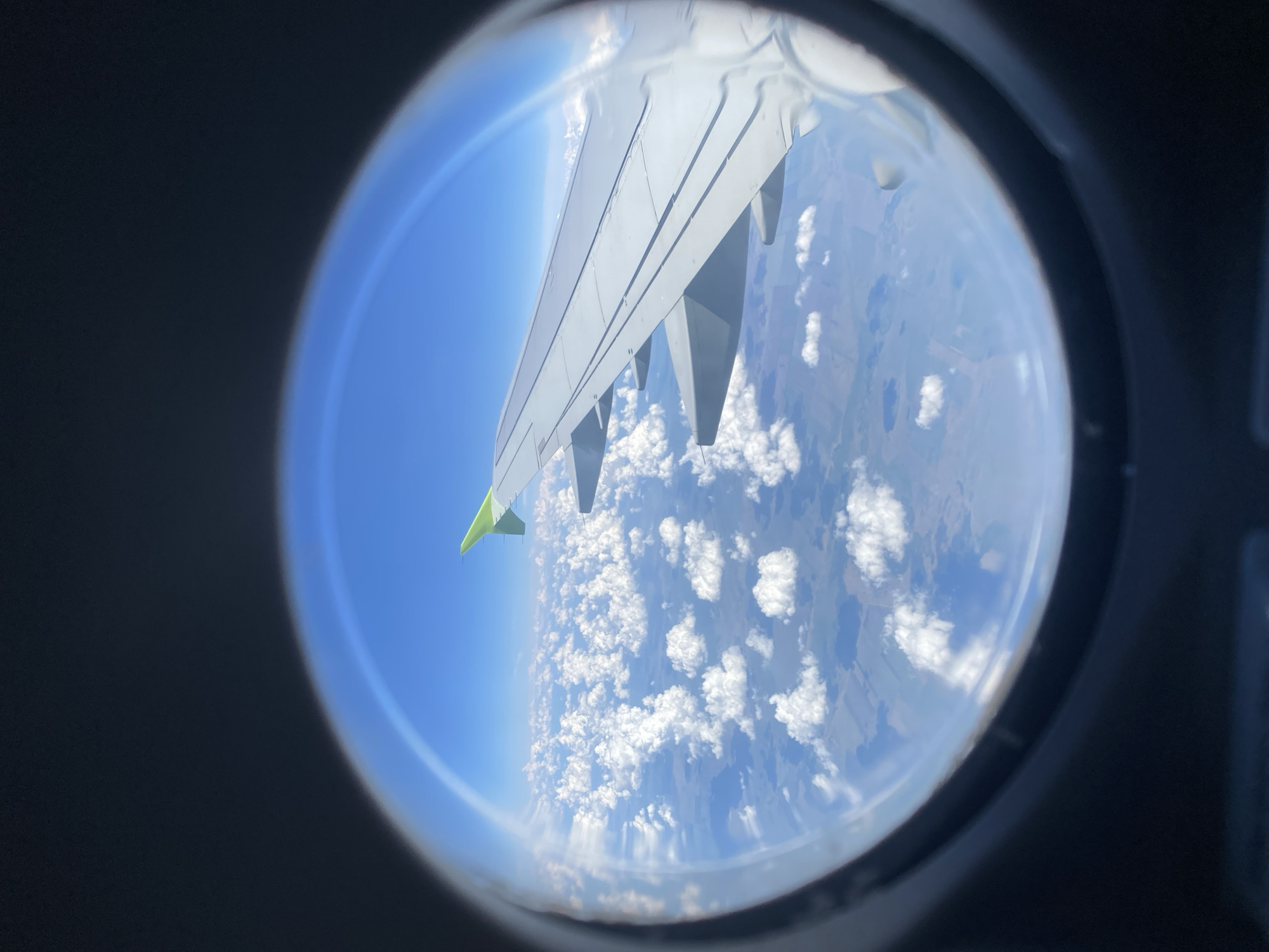 The image shows a view from an airplane window, looking down at the earth from above. The main subjects are the wing of the airplane and the landscape below. The wing, which is light gray with a green tip, extends from the left side of the frame. Below the wing, a vast expanse of sky is visible, dotted with fluffy white clouds. The land below appears as a patchwork of fields and possibly some bodies of water, viewed through gaps in the clouds. The bright blue sky suggests it is daytime, likely mid-morning or afternoon, with clear weather conditions above the cloud layer. The perspective of looking down from a high altitude gives a sense of depth and distance. The dark frame of the airplane window surrounds the view, creating a circular framing effect. There is no visible text in the image. The setting is a flight, likely over a rural area based on the visible land features.
