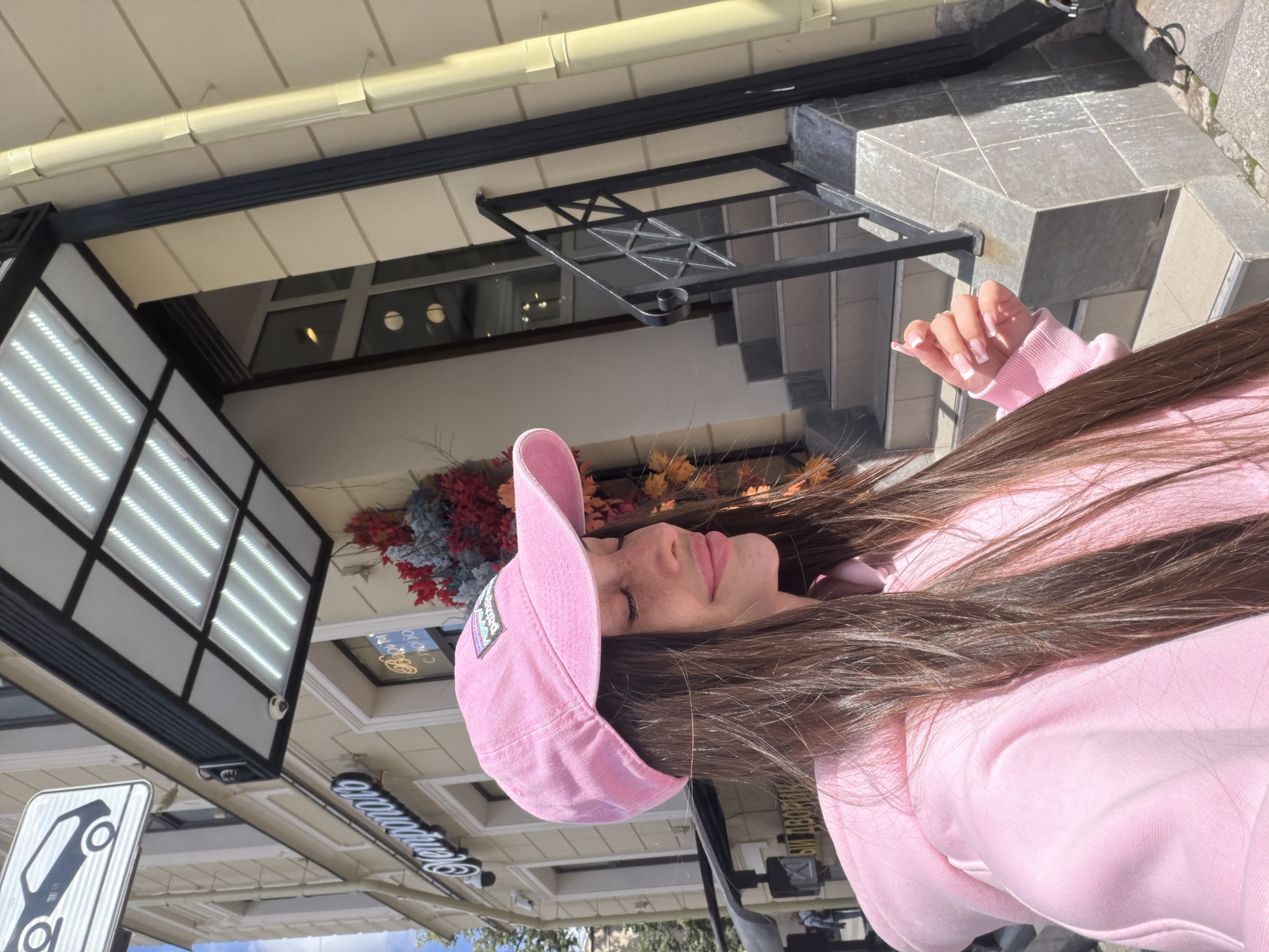 A young woman with long, dark brown hair and a pink baseball cap is posing for a selfie outdoors in front of a building. She is wearing a pink hooded sweatshirt. Her eyes are closed, and she is smiling slightly. The building behind her has a light yellow facade and a dark awning with lights. There is a black railing and stairs leading up to an entrance. A sign with a silhouette of a car is visible above the awning. The scene appears to be in a city, with pedestrian traffic indicated by a figure in the background. The environment suggests a daytime setting with sunlight. The building facade features a sign with Cyrillic lettering that reads "Националь," suggesting the location is likely Saint Petersburg, Russia, possibly near the National Hotel.