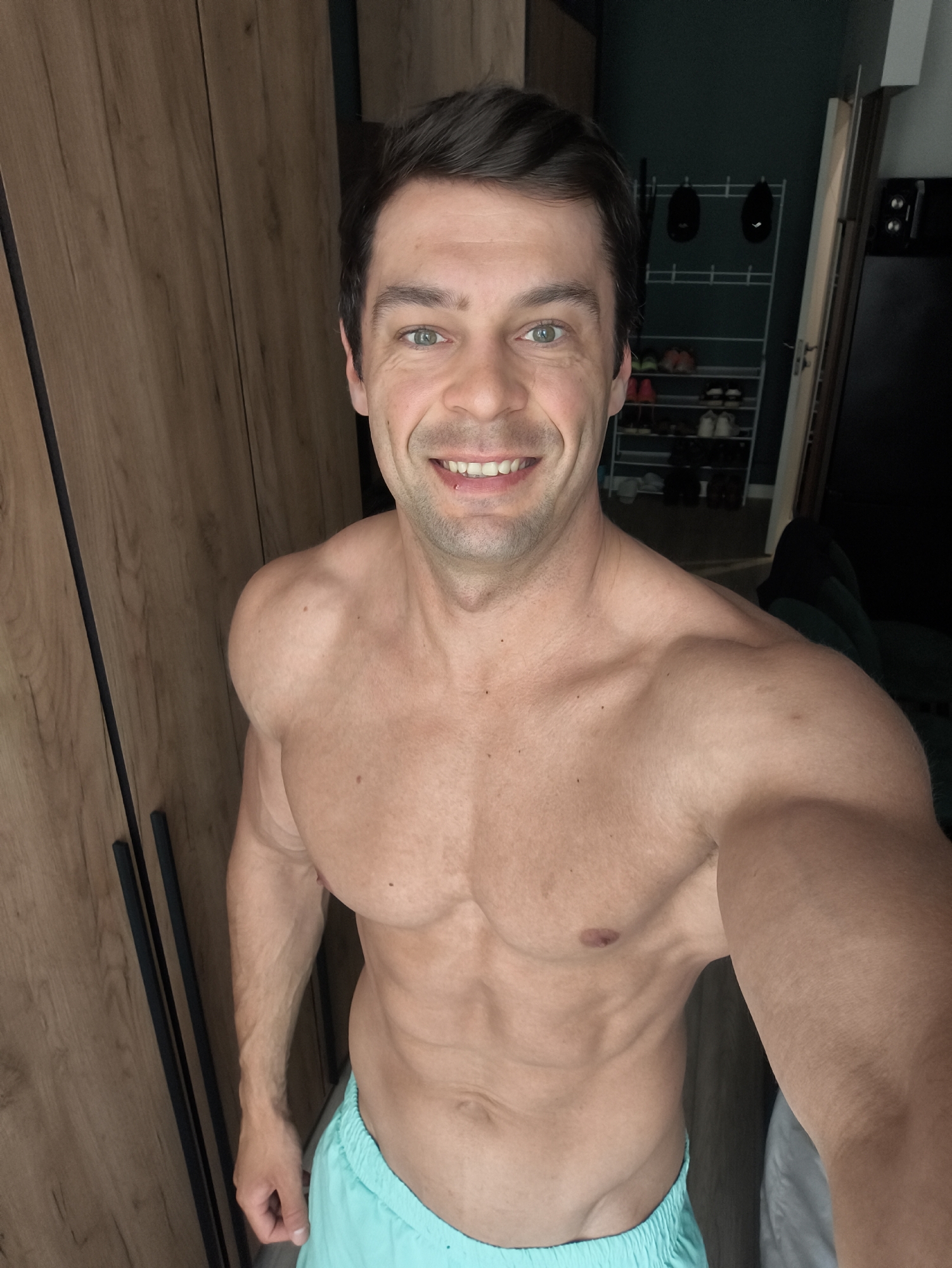 A shirtless man with short dark hair and green eyes is smiling at the camera. He appears to be in a room with a light-brown wooden wardrobe on the left and a rack with shoes and hats in the background. He is wearing turquoise shorts. The location is not discernible as Moscow, Russia based on the given image.