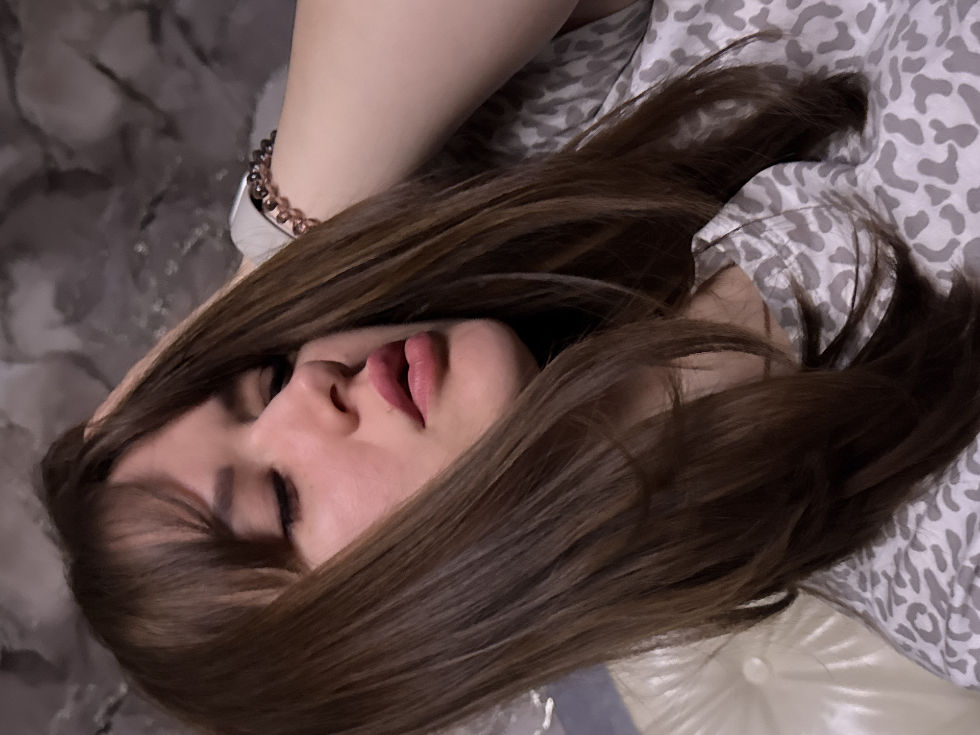 A woman with long, brown hair is lying down, facing slightly to the left. Her eyes are closed, and her lips are slightly parted. She wears a bracelet on her left wrist, and her right arm is visible above her head. The background consists of patterned bedding in shades of grey and white. The location is the city of Chaikovsky, Russia.