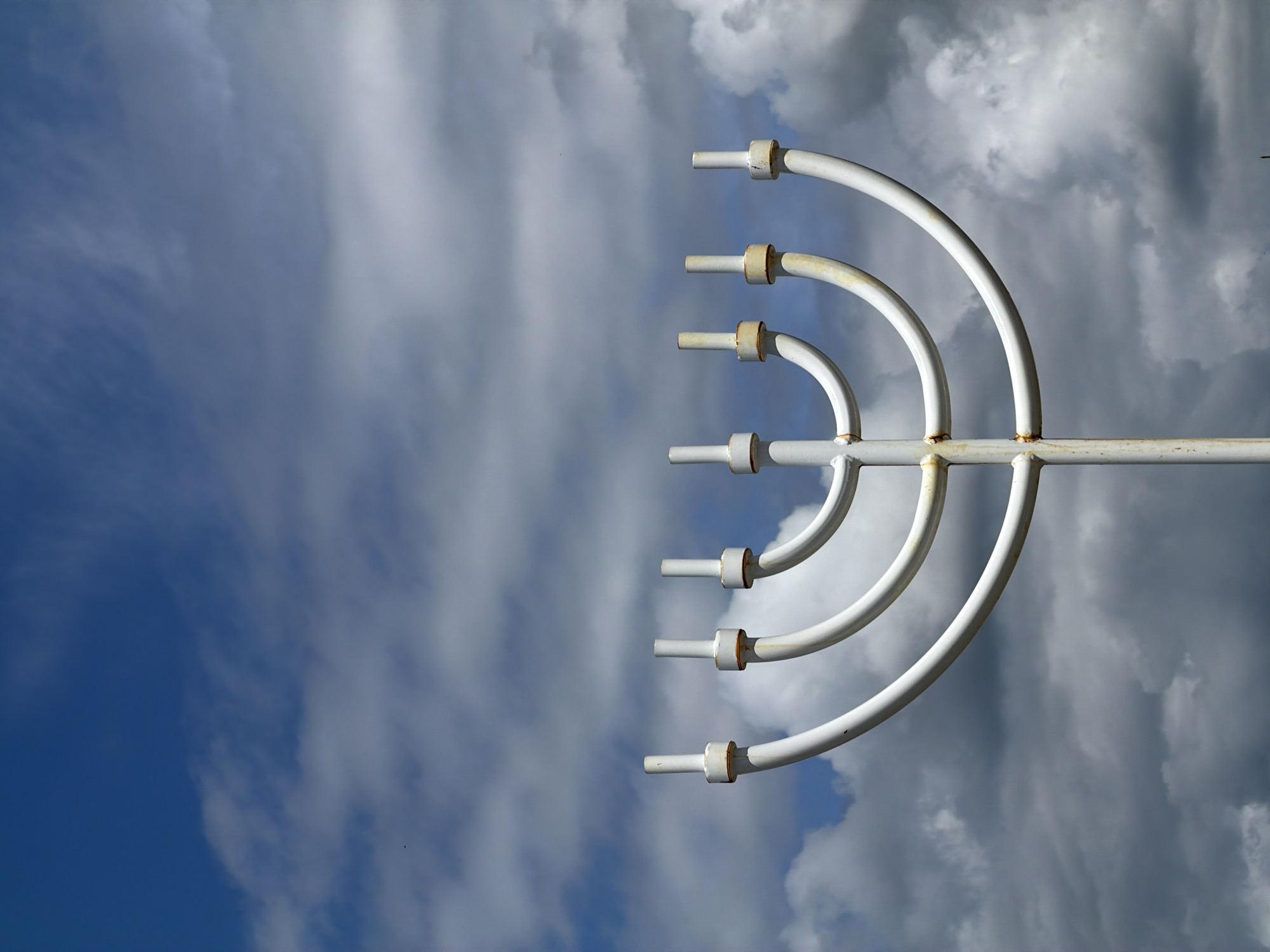 A white metal menorah stands against a backdrop of a partly cloudy blue sky. The menorah is a large, outdoor sculpture with seven branches, each ending in a cylindrical candle holder. Some of the holders show slight signs of rust. The central stem of the menorah is also visible. The sky above is a mix of bright blue and darker, textured clouds. The image appears to be taken from a low angle, looking up at the menorah. This menorah is located in Birobidzhan, Russia, serving as a prominent symbol in the city.