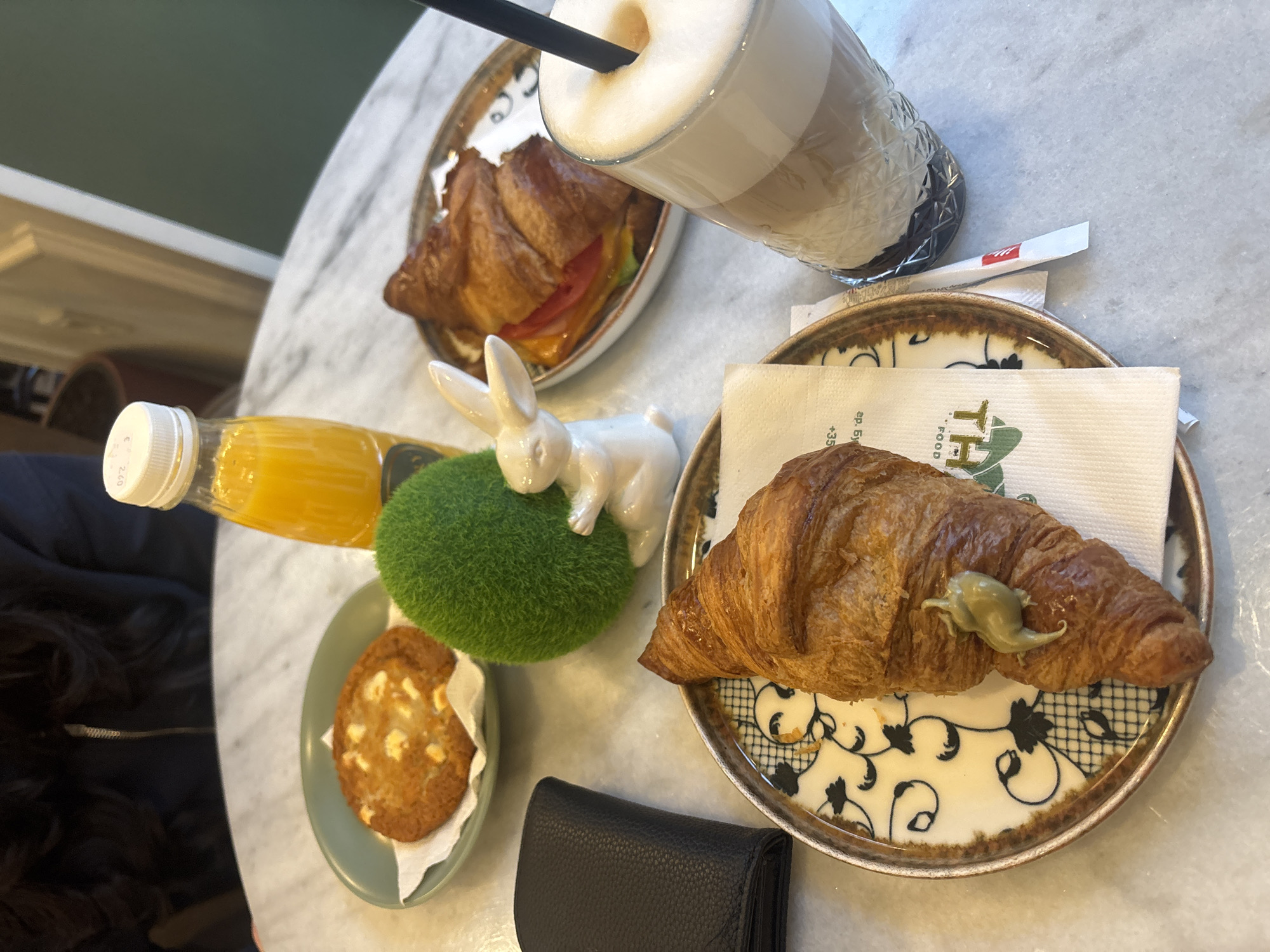 The image depicts a table setting in Burgas, Bulgaria, featuring a latte with a black straw, a croissant sandwich, a single croissant with a green topping, a cookie, a bottle of orange juice, a small ceramic rabbit figurine on a green decorative ball, a sugar packet, and a black wallet. All items are arranged on a light-colored marble-patterned table.