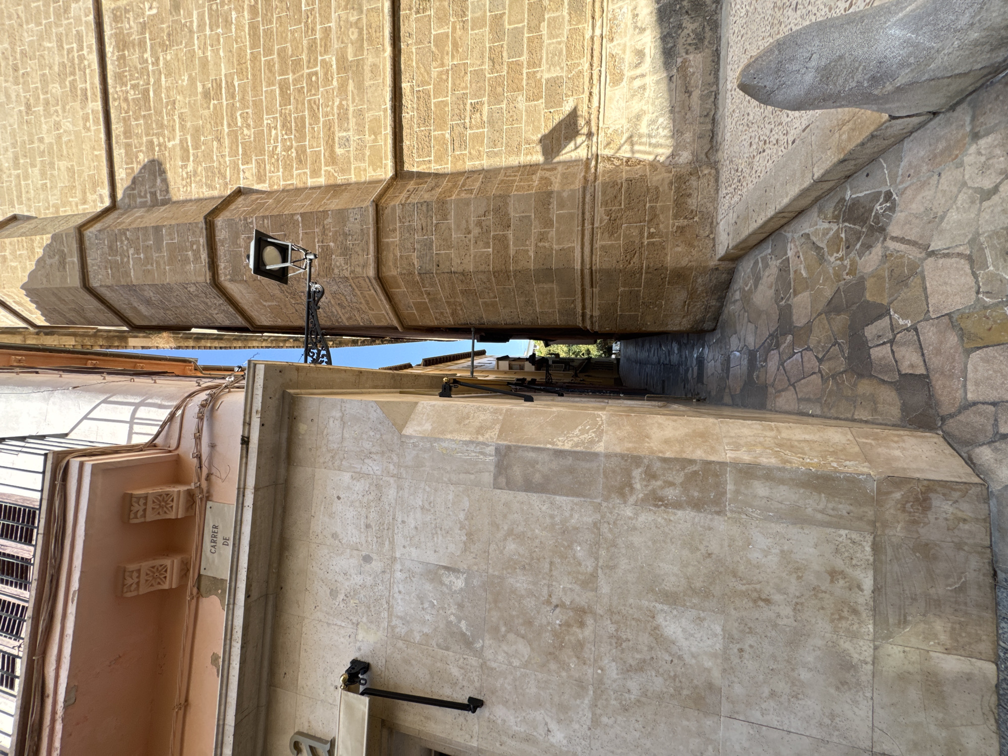 The image shows a narrow alleyway in Palma de Mallorca, Spain. On the left, a building with a pink facade and decorative brackets is visible. A street sign reading "CARRER DE" is affixed to the wall. The building's wall is made of large, light-colored stone tiles. On the right, a tall structure constructed of tan brick dominates the scene. A black wrought-iron lantern is mounted on the brick building. The alleyway itself is paved with irregular flagstones, and a curved, grey stone object juts out into the foreground from the right. Sunlight casts shadows from the buildings and the lantern onto the brick wall and the street. The alleyway recedes into the distance, where glimpses of green foliage and another building can be seen.