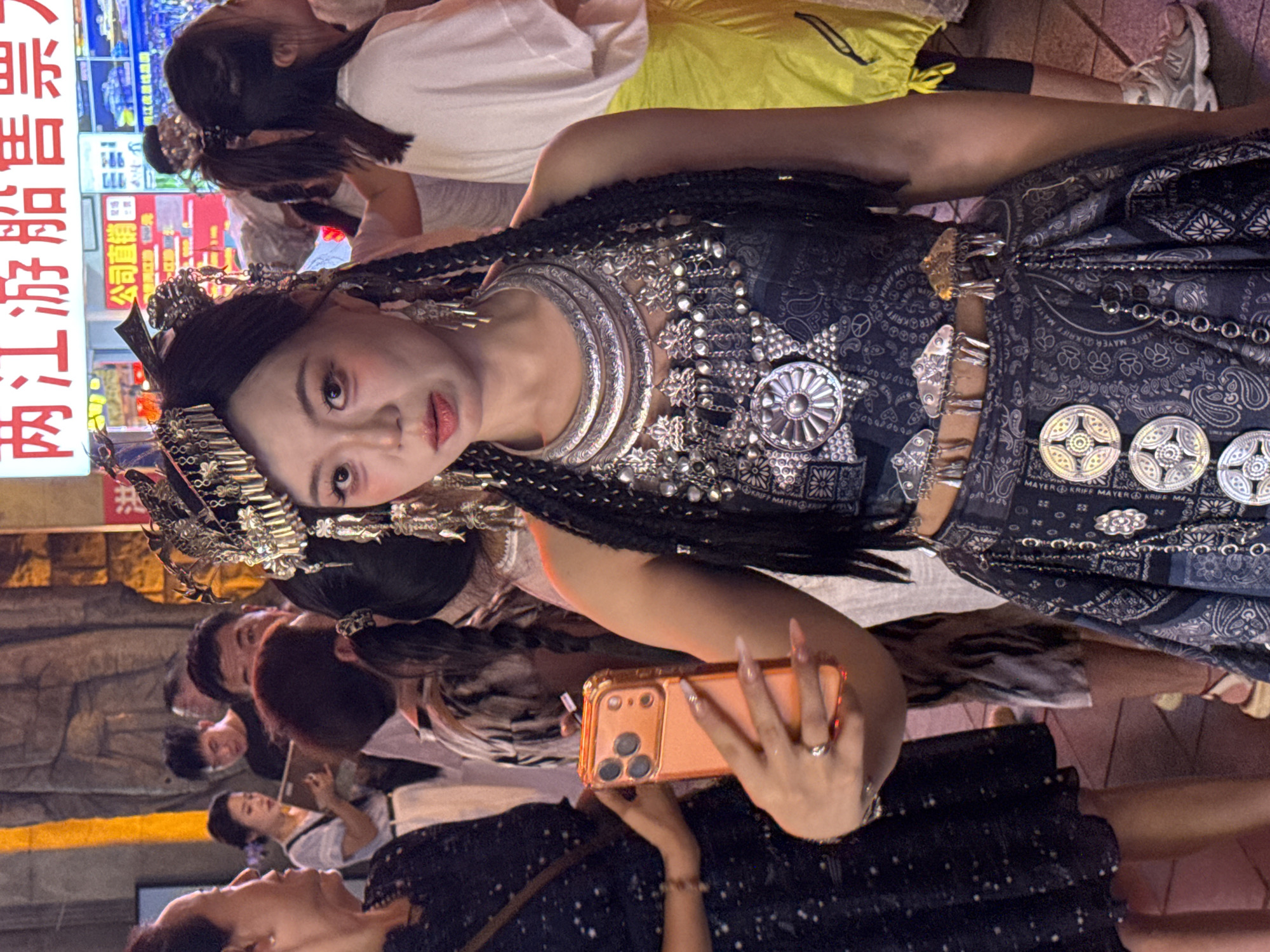 In Yuzhong District, China, a young woman with dark hair styled in braids and adorned with intricate silver headwear and jewelry, including a large, layered necklace and matching earrings, poses for a selfie. She is wearing a dark, patterned outfit with silver accents. She holds an orange smartphone in her right hand, angled towards herself. Behind her, a crowd of people are visible, some looking towards the camera and others engaged in their own activities. The background suggests an outdoor setting, possibly a street or market, with illuminated signs in Chinese characters, one of which reads "两江游船售票" (Liangjiang Cruise Ticket Sales). The lighting is dim, indicating it is likely evening or nighttime.