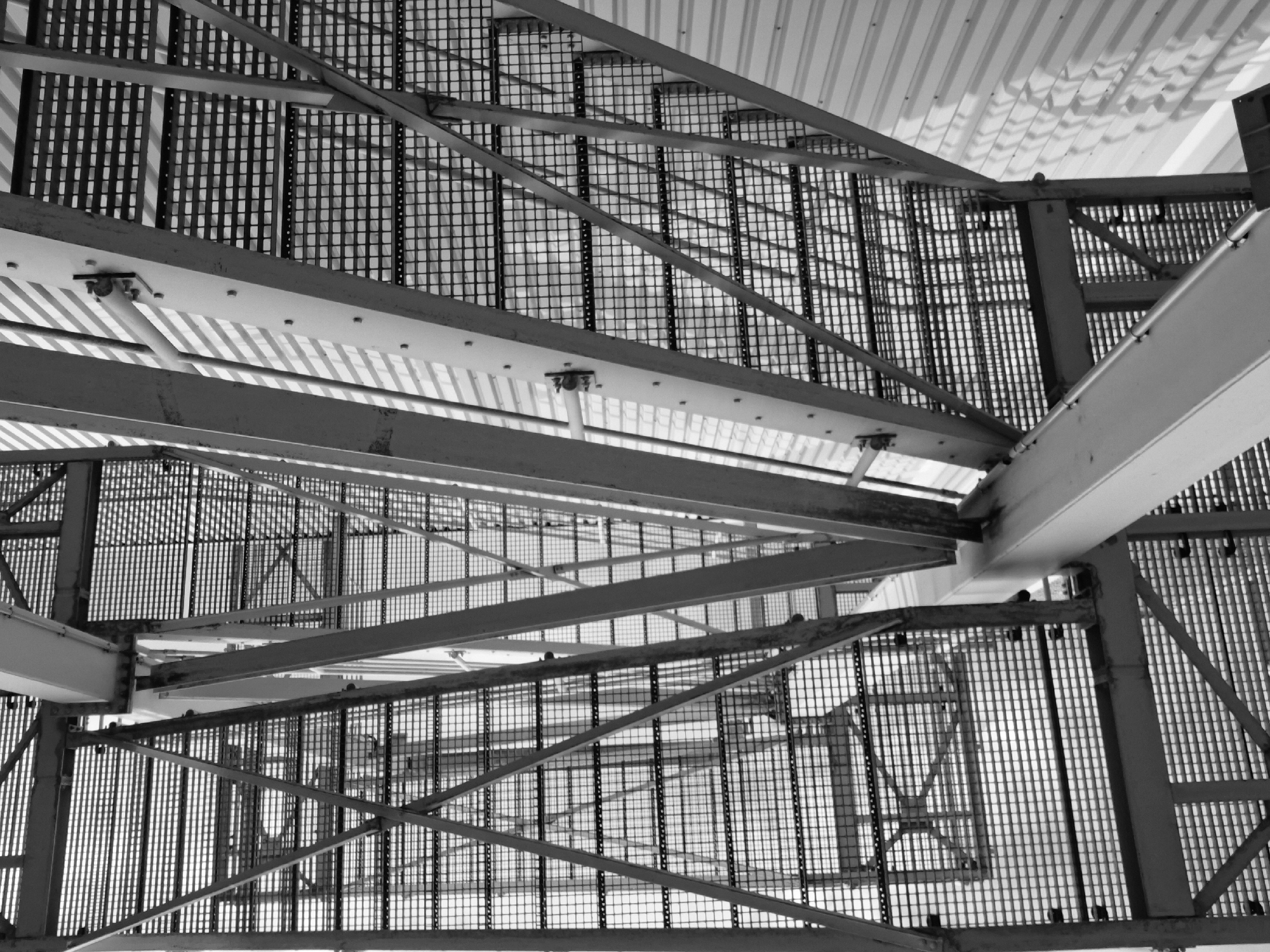 This is a black and white, low-angle, close-up shot of a metal staircase structure, likely an industrial or architectural element, as seen from below. The staircase is constructed with grated metal steps and reinforced with metal beams and supports forming a complex geometric pattern. The background consists of a corrugated metal wall with a repeating ridged pattern, casting striped shadows across the structure. The lighting suggests an overcast day or diffused artificial light, with sharp contrasts between the metallic surfaces and the shadows. The image does not contain any people or identifiable landmarks that would place it specifically within the city of Rastatt, Germany.