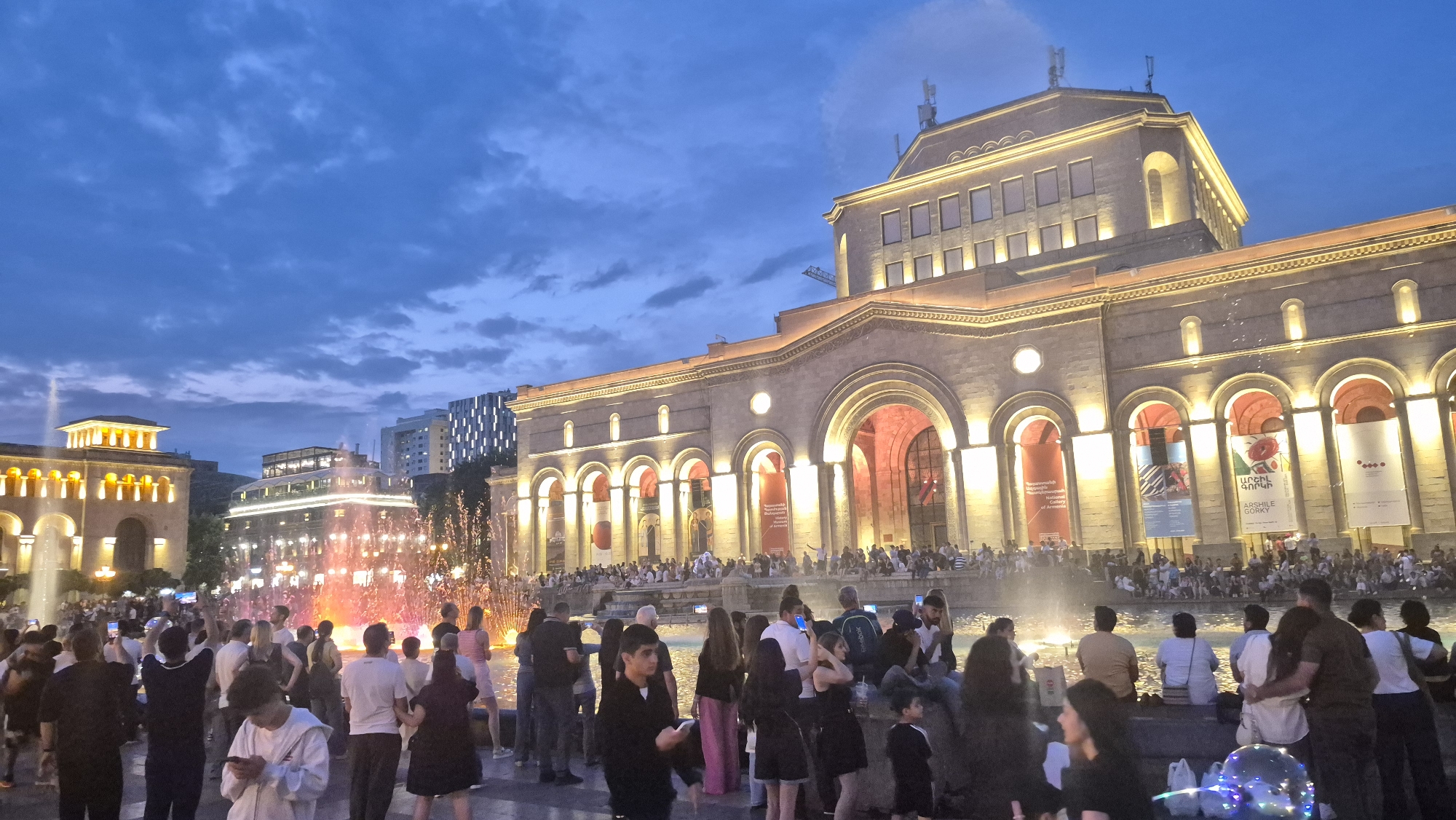 A wide-angle shot captures an evening scene in a city square, featuring a grand, illuminated stone building with arched entrances and classical architectural elements. The building's facade is lit with warm yellow lights, highlighting its imposing structure against a darkening blue sky with scattered clouds. In the foreground, a lively crowd of people are gathered around a brightly lit fountain, its water illuminated with vibrant orange and pink hues. Some individuals are holding up phones, possibly taking pictures or videos. The atmosphere suggests a public gathering or event. To the left of the main building, another structure with similar architectural features is also illuminated. In the background, modern buildings with geometric patterns of lights are visible, contrasting with the historical architecture. The overall scene depicts a vibrant urban environment with a blend of historical and modern elements, set during twilight.