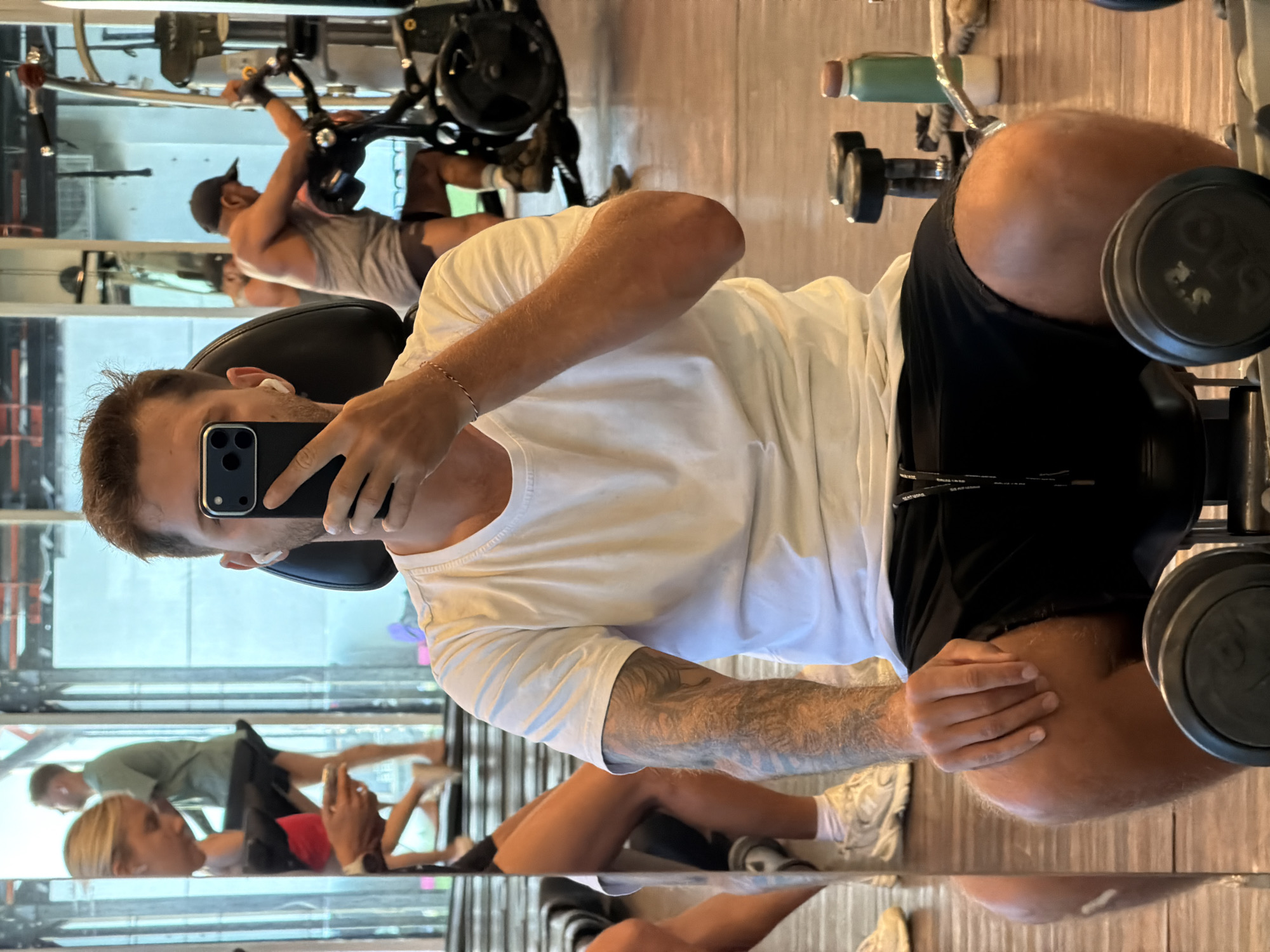 The image is a vertical mirror selfie taken by a man in a modern gym, located in North Kuta, Indonesia. The main subject, positioned in the foreground, is wearing a white t-shirt and black shorts, with a white earbud in his right ear. He holds a black iPhone with three camera lenses, mostly obscuring his face as he takes the photo. A subtle bracelet is visible on his left wrist, and tattoos adorn his arm.
The reflections in the mirrored walls reveal a busy gym environment. In the background, another man, dressed in a dark cap and light tank top, is actively using a leg press machine. Further down and to the left in the reflection, a blonde woman is seen lying on what appears to be a bench, focused on her smartphone. Another man in a green t-shirt is faintly visible in the deeper reflection.
The gym features mirrored walls that create multiple reflections and a light wooden floor. Various pieces of gym equipment are scattered throughout the background, including dumbbells, one of which shows the number "2.5" on its plate, and a green and white water bottle on a shelf. Partially visible text on the main subject's black shorts appears to read "SKATEBOARDING". The bright, even lighting suggests a daytime setting. The scene depicts a typical, active workout session in a fitness center.