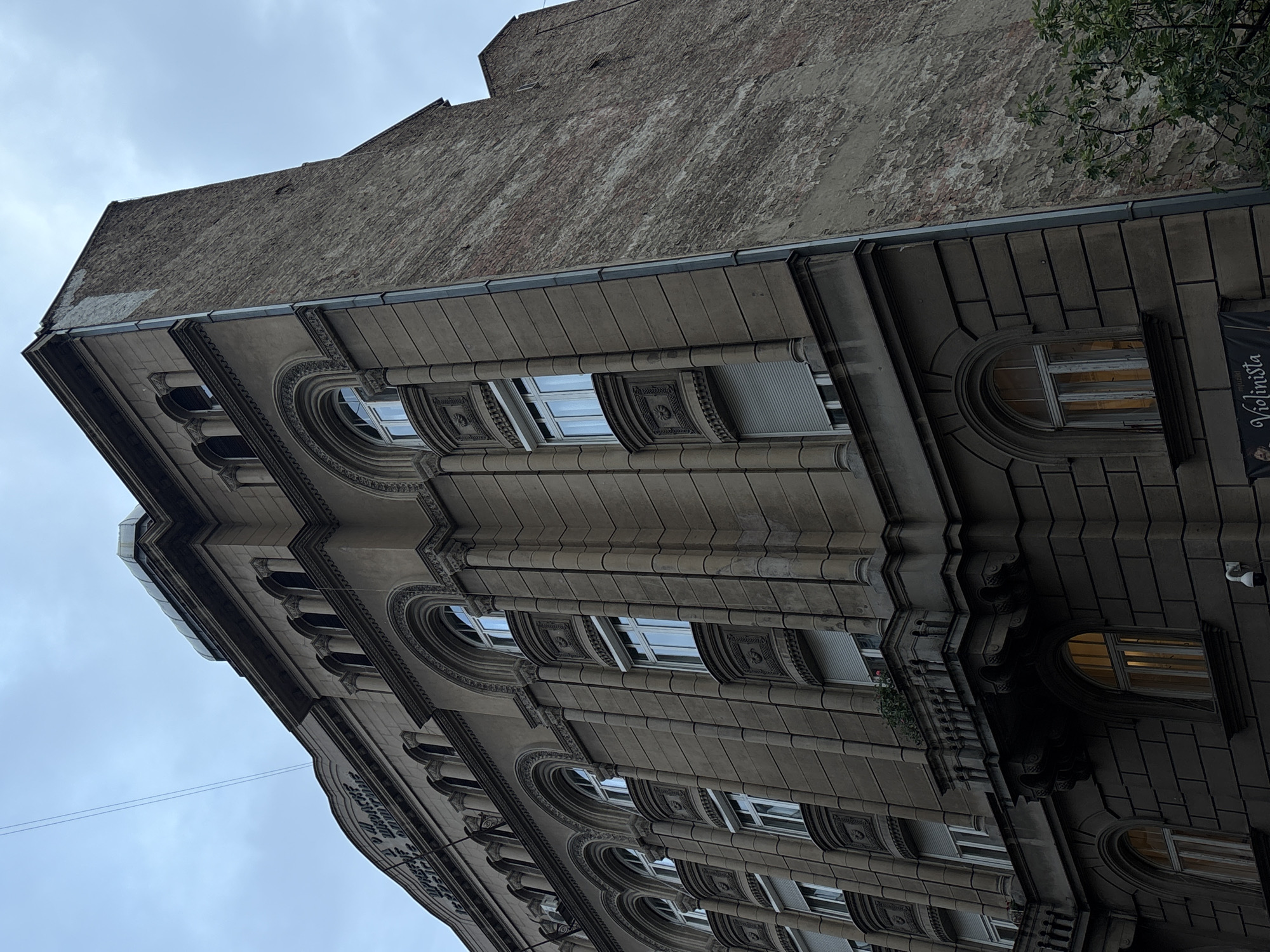 The image depicts the upward view of a multi-story building in Belgrade, Serbia. The architectural style is classic European, with rounded windows, ornamental facades, and a gray stone or brick exterior. The building's upper section appears aged with worn or patched stonework. A few windows are visible with glimpses of blinds or interiors. A sign labeled "Violinista" is attached to the side of the building. The sky is overcast.