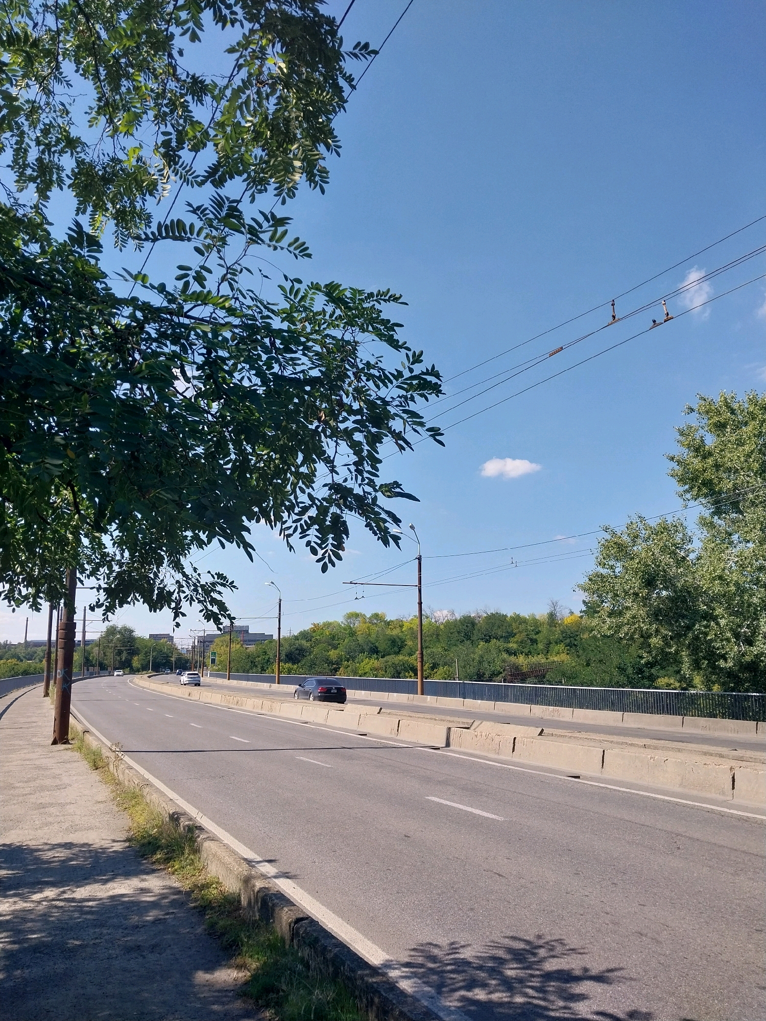 The image depicts a daytime view of an elevated road, likely a bridge, in Dnipro, Ukraine. A sidewalk runs along the left side of the road, shaded by the leaves of a large tree. The road itself is asphalt with white lane markings and appears to have at least two lanes in each direction, separated by concrete barriers. Several cars are visible driving on the road. Beyond the road, a dense line of green trees forms a natural embankment, and in the distance, some buildings can be seen. The sky is clear blue with a few wispy clouds. Overhead, power lines stretch across the frame, supported by utility poles. The scene is devoid of any individuals in the immediate foreground. The overall impression is that of a typical urban thoroughfare on a sunny day.