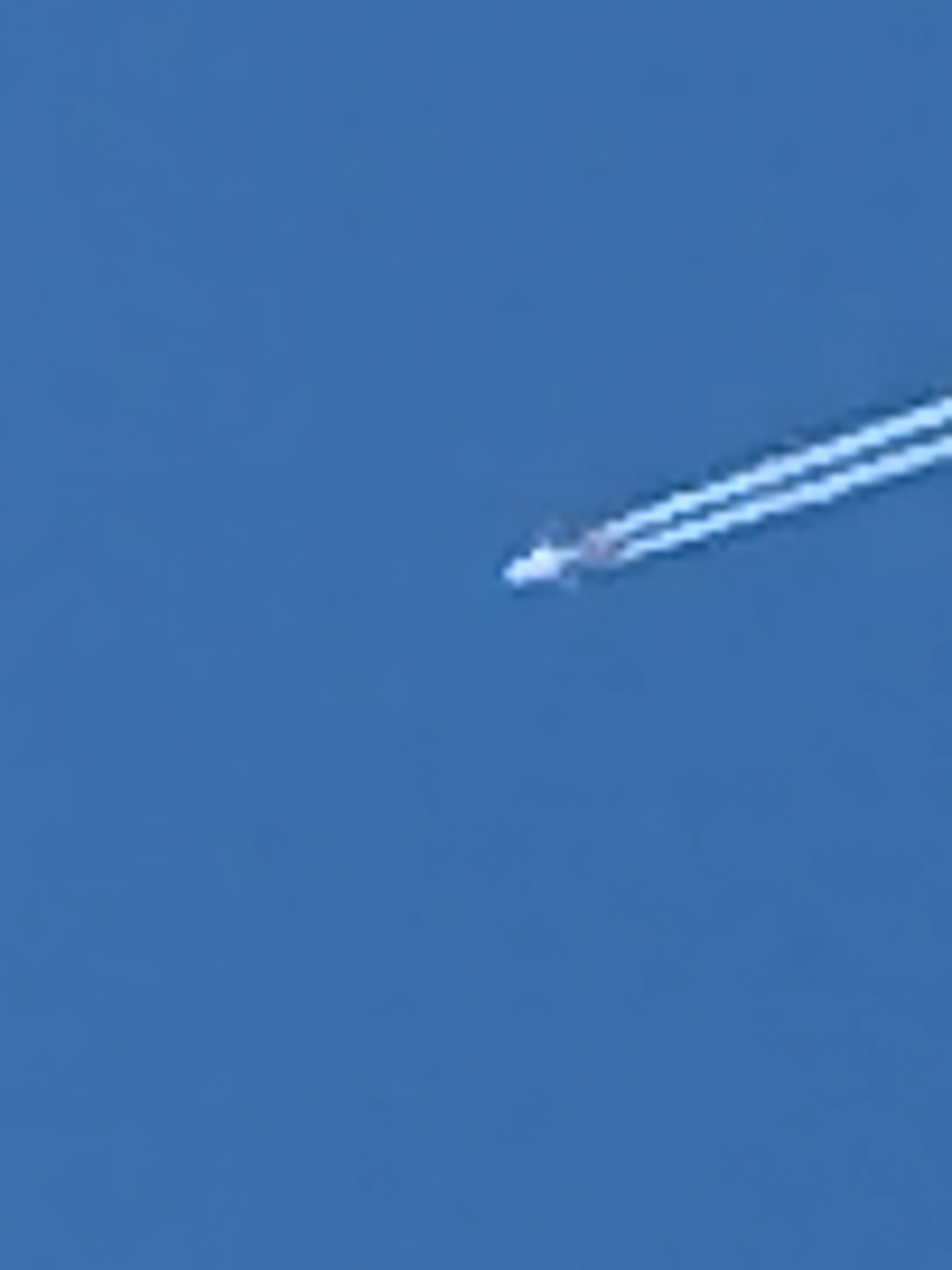 An airplane is seen leaving a contrail across a clear blue sky. The airplane itself is a small, white object at the front of the contrail, which consists of two parallel white lines that stretch diagonally from the upper right to the lower left of the image. The background is a uniform, bright blue, indicating it is daytime with no clouds.