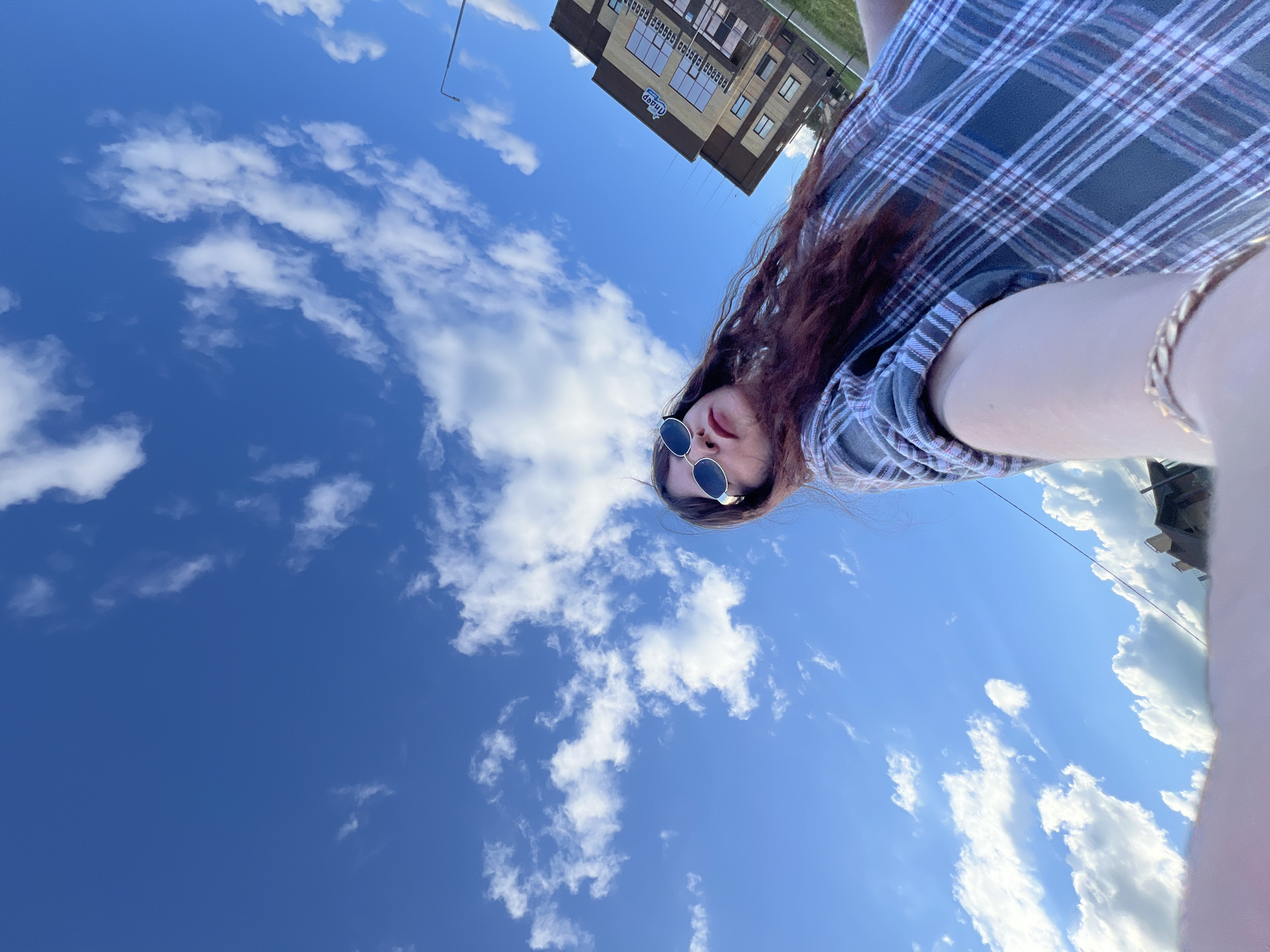 An individual is pictured in this low-angle, outdoor selfie. They are wearing a plaid shirt and sunglasses, with long, dark hair visible. The background is a bright blue sky with scattered white clouds, and a multi-story building with a sign reading "Anapaer" is visible to the right, suggesting the location is Noviy, Russia. A lamppost stands near the building. The image appears to be taken during daylight hours.