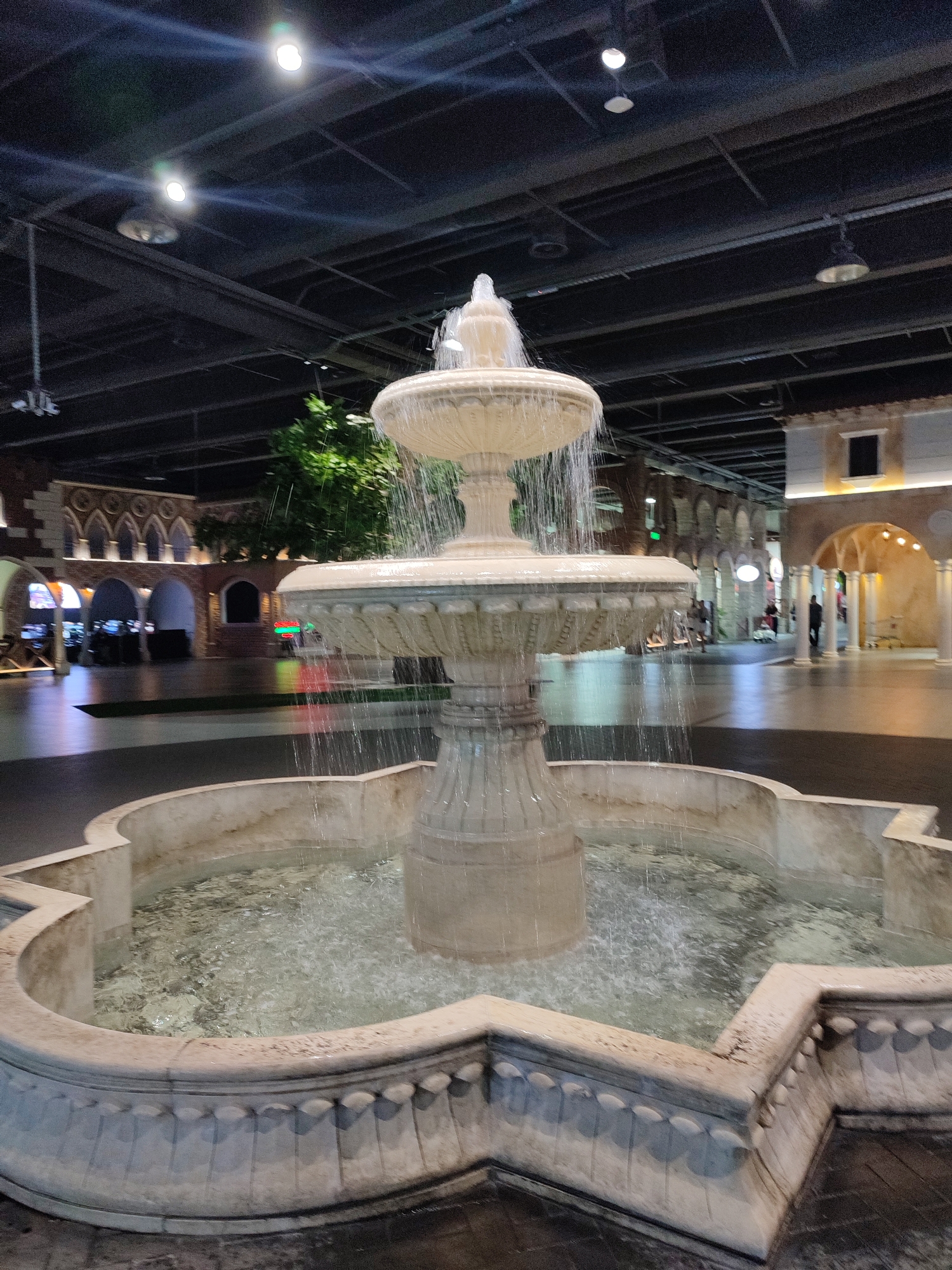 The image depicts a multi-tiered fountain in the foreground, with water cascading down its ornate stone structure. The fountain is located within a brightly lit indoor space, possibly a shopping mall or entertainment complex, given the architectural style resembling a European old town. The background reveals colonnades, arched walkways, and building facades with detailed stonework. Artificial trees and indoor lighting suggest an indoor environment designed to mimic an outdoor setting.
Several people are visible in the background, appearing to be shoppers or visitors, some walking and others standing. Their presence suggests the space is open and active. The lighting in the scene is artificial, with bright overhead lights illuminating the area, casting reflections on the wet floor around the fountain. The time of day cannot be determined from the image, but the indoor setting implies it could be any time, day or night. The weather is irrelevant as it is an indoor environment.
Notable details include the decorative carvings on the fountain and the architectural elements in the background, such as the arches and cornices, which evoke a sense of classical or Mediterranean design. The reflective surface of the floor around the fountain indicates it is wet from the water spray. There is no visible text in the image. The overall impression is one of a well-maintained, aesthetically pleasing public space designed for leisure or commerce.