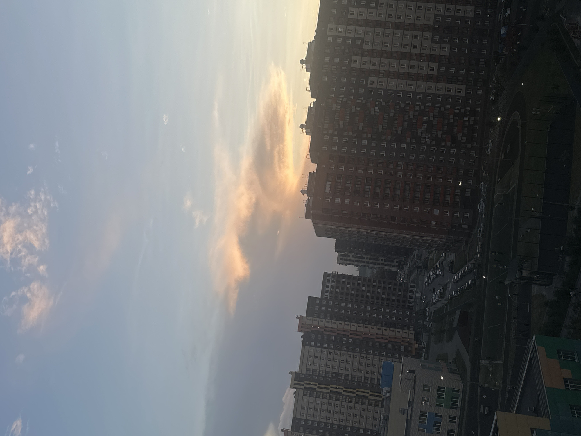 The image depicts an aerial view of a residential area in Mirny, Russia, during what appears to be either sunrise or sunset. The sky is a gradient of pale blue, transitioning to hues of pink and orange where clouds are present. Dominating the scene are several multi-story apartment buildings, characterized by their uniform window patterns and varying architectural styles. Some buildings feature light-colored facades with darker window frames, while others have more colorful exteriors with balconies.
In the foreground and midground, there are paved areas with numerous parked cars, indicating a densely populated urban environment. Adjacent to some buildings are sports fields, possibly soccer fields, with visible markings and fencing. A few individuals are sparsely scattered across these fields, engaged in activities that are indistinct due to the distance and lighting. One building in the lower left of the frame appears to be a school or community center, with a distinct multi-colored facade featuring shades of blue, green, and yellow. The overall impression is a modern, albeit somewhat uniform, residential development within a city.