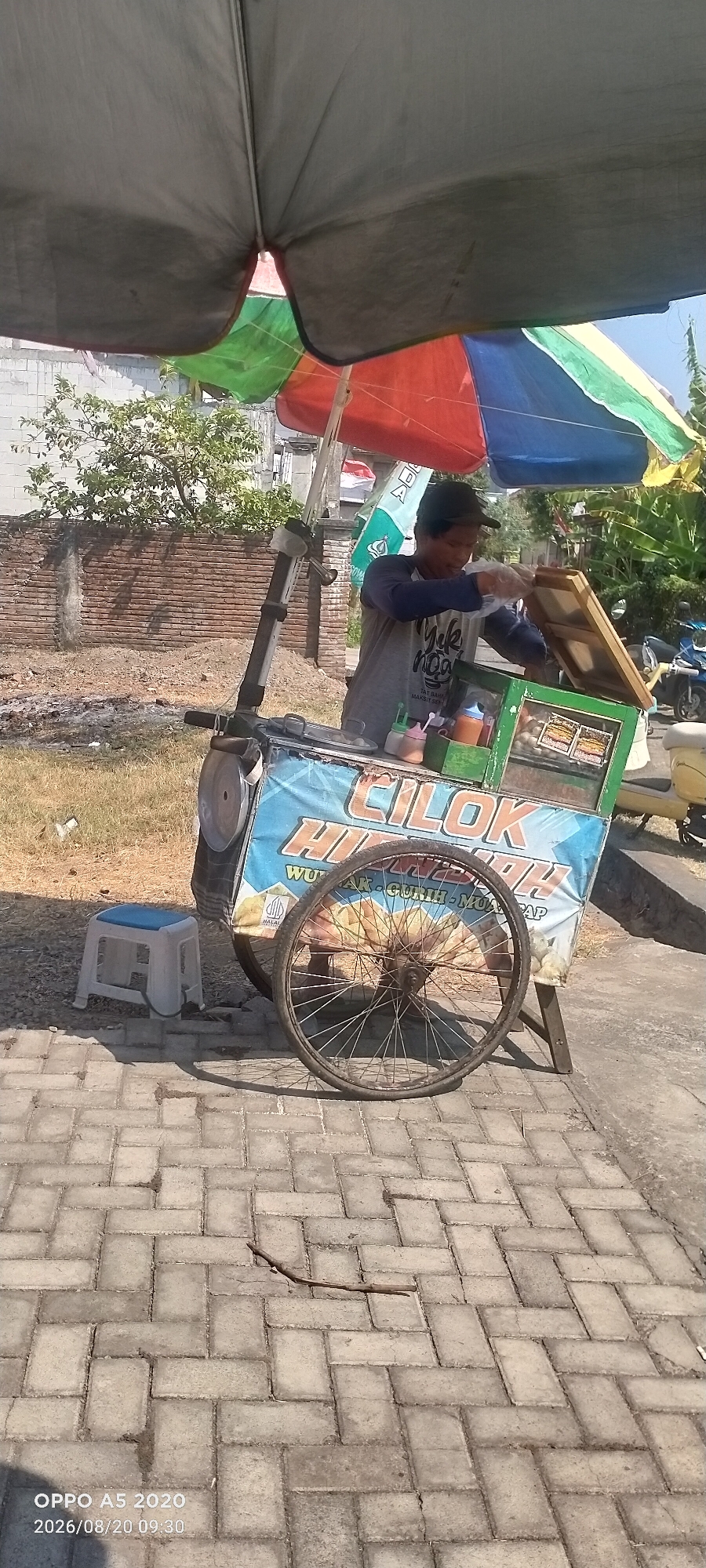 A street vendor is serving food from a cart under a colorful umbrella on a paved walkway in Sowan Kidul, Indonesia. The vendor, wearing a grey t-shirt and a dark cap, is handing a plastic-wrapped item to a customer whose hands are not visible. The cart displays the text "CILOK HI....", indicating the type of food being sold, likely "cilok," a traditional Indonesian snack. Several bottles of sauce are visible on the cart. A white plastic stool is placed to the left of the cart. The background shows a brick wall, dry grass, trees, and a motorcycle. The sun is bright, casting shadows on the ground. The timestamp "OPPO A5 2020 2026/08/20 09:30" is visible in the lower left corner.