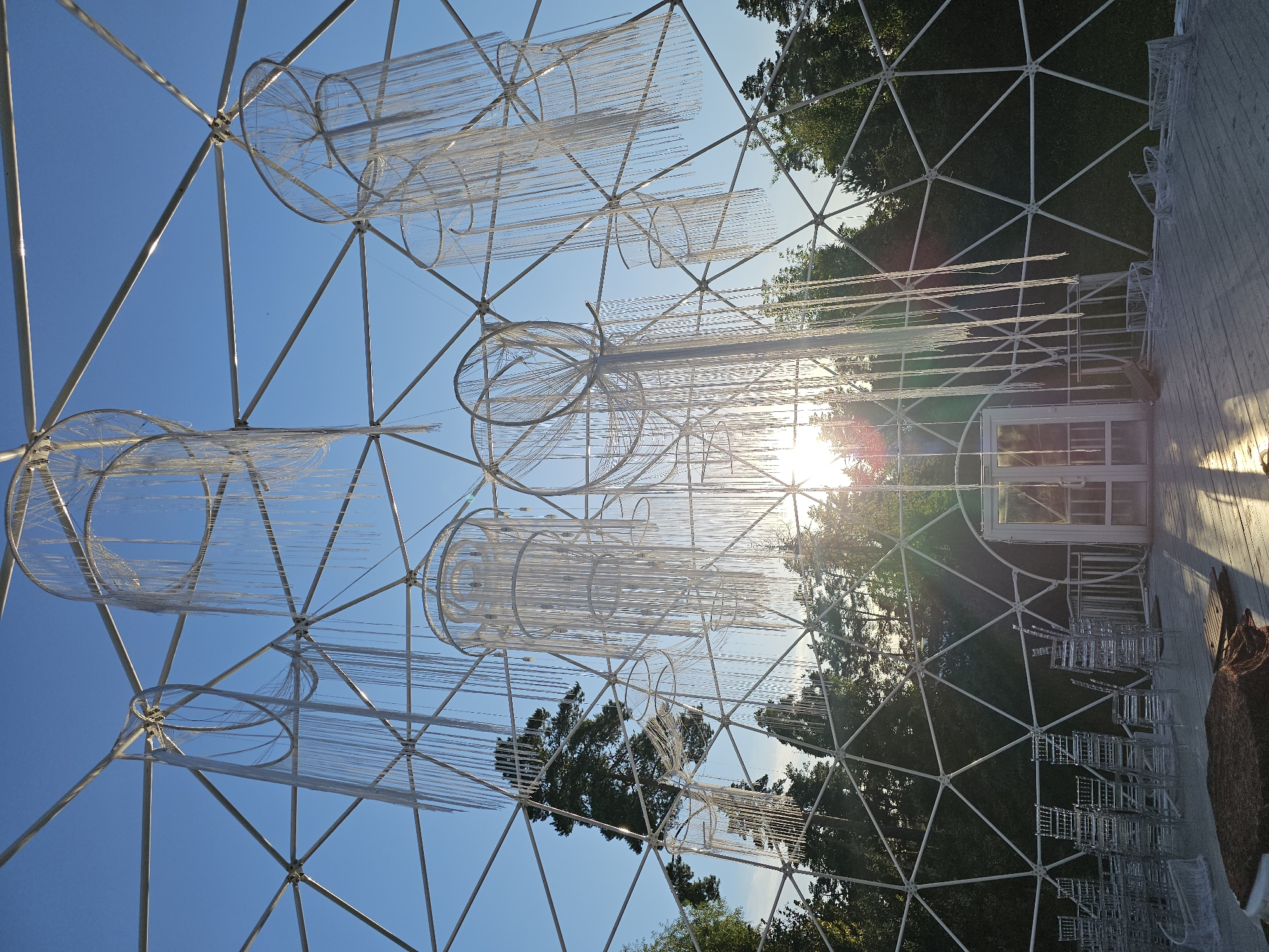 The image shows the interior of a geodesic dome structure with a white metal frame against a bright blue sky. Inside the dome, several large, cylindrical chandeliers made of white hanging strands are suspended from the frame. The sun is setting, casting a bright glare through the dome and illuminating the interior. In the foreground, several stacks of clear plastic chairs are arranged on a wooden floor. A glass door with a white frame is visible in the background. Trees are seen outside the dome, silhouetted against the setting sun. The location is identified as Ulitkino, Russia.
