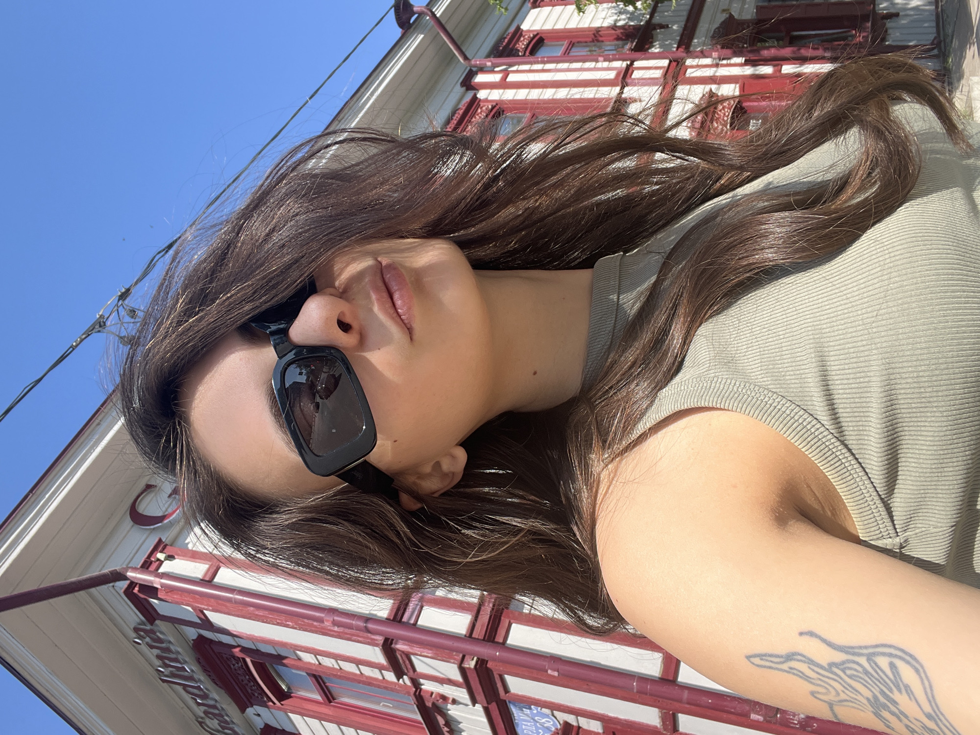 A young woman with long, brown hair and sunglasses poses for a selfie outdoors. She is wearing a sleeveless, olive-green ribbed top. Behind her is a red and white building with ornate trim and several windows. The building appears to be a historical structure. The sky is clear and blue. A tattoo of a flame-like design is visible on her left arm. The image was taken on a sunny day. The building in the background has "Giardiniia" written in red lettering across its facade. A partial sign with Cyrillic letters and the number "38" is visible on the building. This location is likely in the city of Krasnoyarsk, Russia.