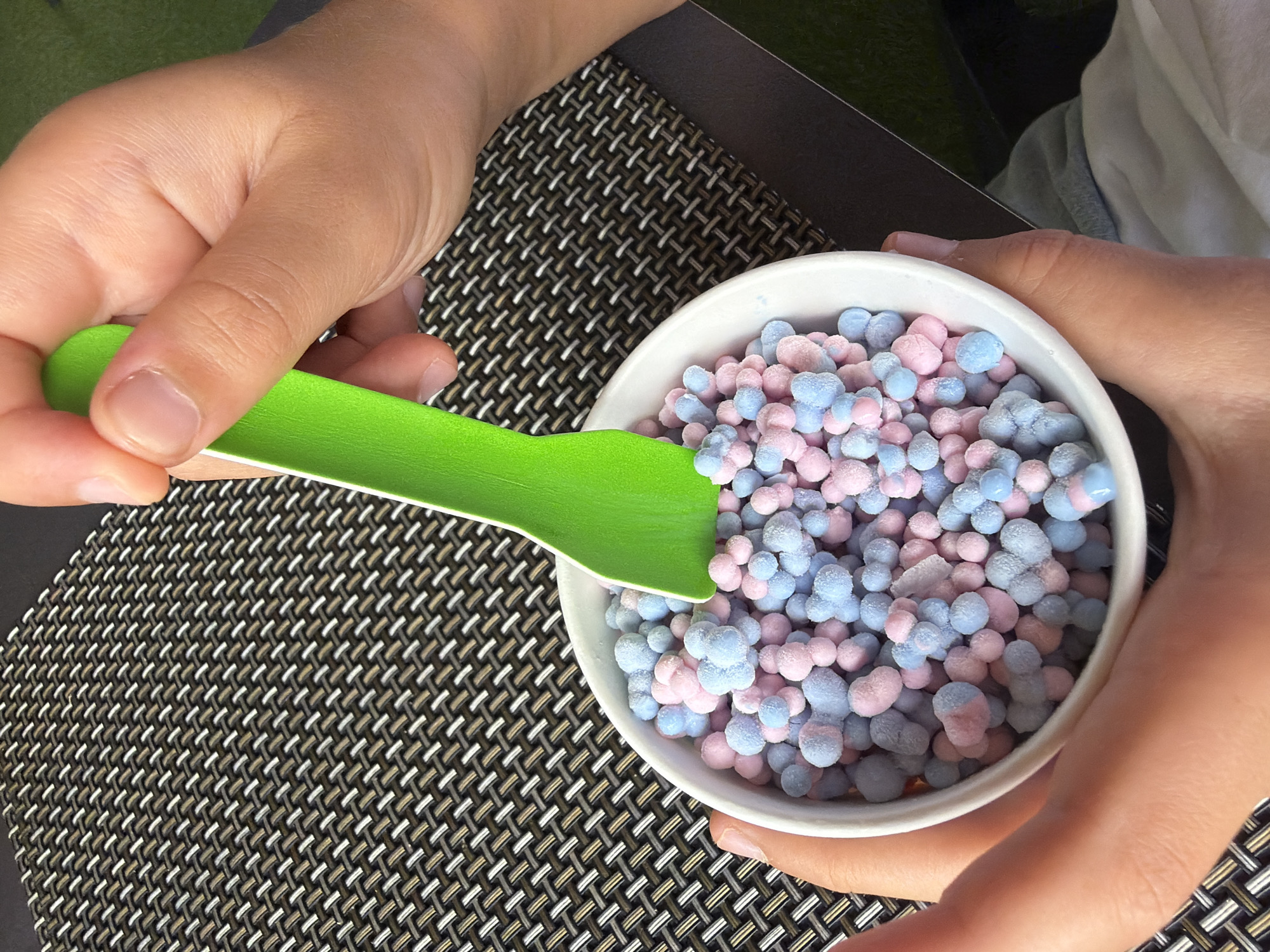The image shows a close-up of a white bowl filled with small, round, pastel-colored pieces of what appears to be ice cream or mochi. The pieces are a mix of light blue and pink, and they are dusted with a light coating of frost. A bright green plastic spoon is partially submerged in the bowl, held by a person's hands. The bowl sits on a textured surface, possibly a placemat, with a pattern of woven brown and white threads. The background is blurred, suggesting an outdoor or casual setting. The person's hands, one holding the bowl and the other holding the spoon, are visible. The lighting is bright, casting soft shadows. There is no information within the image to indicate a specific location in La Vila Joiosa, Spain.
