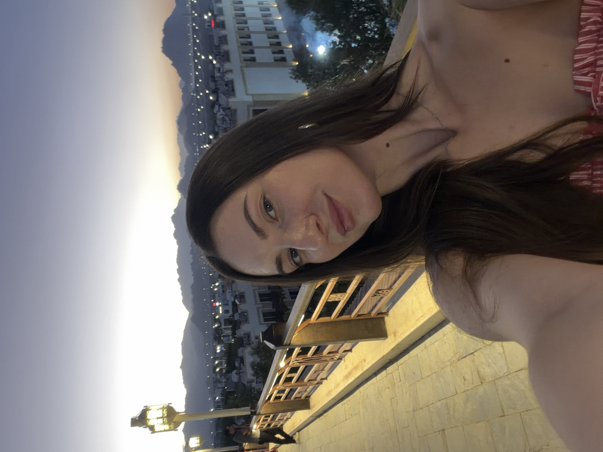 A woman with long, dark hair takes a selfie outdoors at dusk, with a city and mountains in the background. She is wearing a strapless top with red and white stripes and a silver necklace. She has a mole on her neck and another on her chest. Behind her, a person in a gray shirt and dark pants stands near a railing and a lamppost. The railing is made of wood and metal, and the walkway appears to be paved with stone tiles. The city lights are visible in the distance, along with the silhouettes of mountains. The sky is a soft gradient of purple and orange. This scene is likely located in Sharm El Sheikh, Egypt, given the architectural style of the buildings and the mountainous terrain.