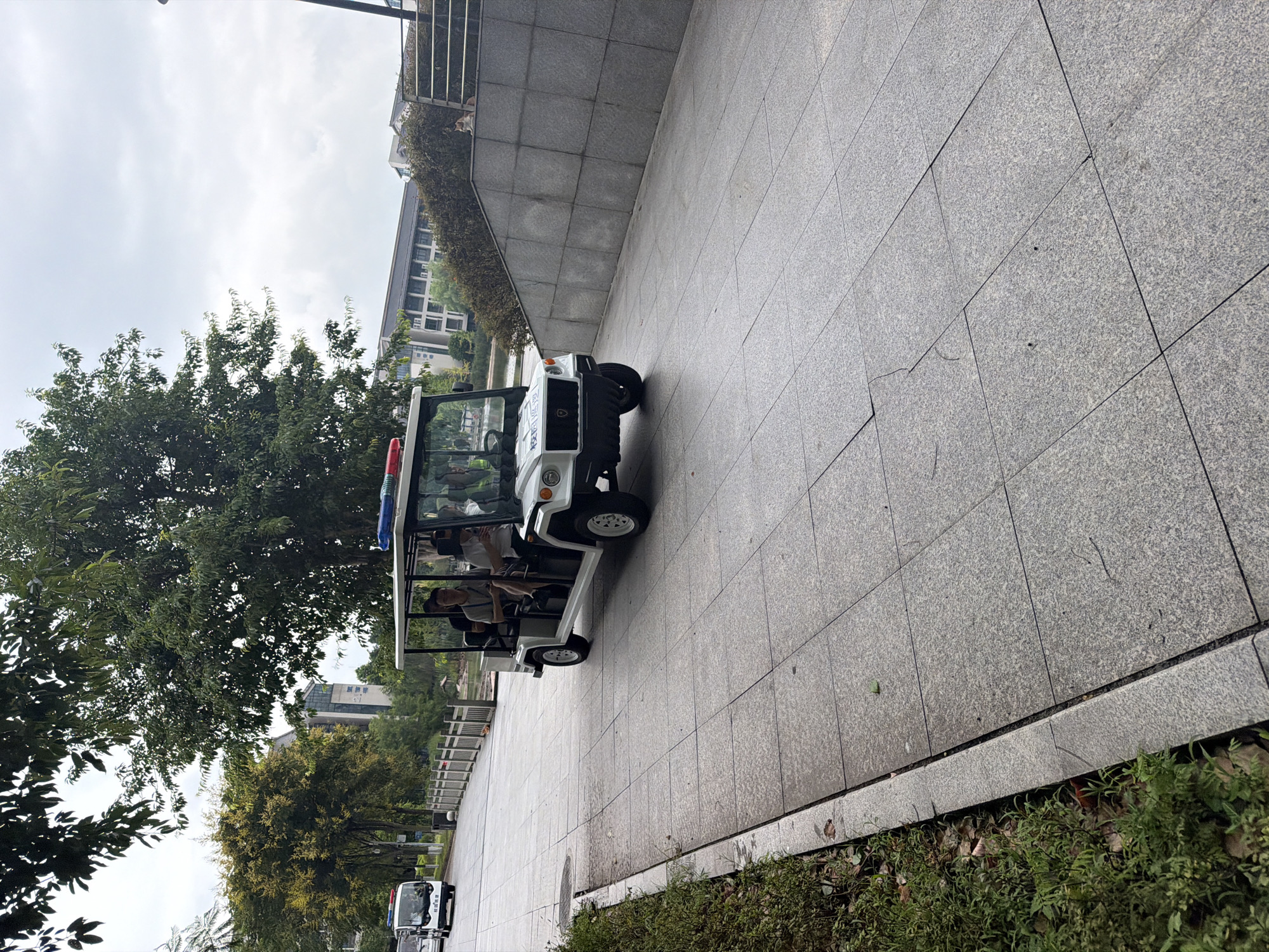 An outdoor, eye-level, medium shot captures a white, open-air electric vehicle with a roof and a light bar containing red and blue lights, resembling a patrol vehicle. The vehicle is on a paved walkway made of large, square, gray tiles. The vehicle is moving forward, and inside, three people are visible. The driver, a man wearing a white shirt, is seated in the front left. To his right, a man in a gray collared shirt is seated. A third person, also a man, is in the back seat. To the left of the electric vehicle, another white vehicle, possibly a mini-van, is further down the walkway. A body of water is visible to the left of the walkway, bordered by a wooden fence. Lush green trees and bushes line the walkway and are situated behind the fence. In the background, modern buildings are visible. The sky is overcast and cloudy. To the right of the walkway, a stone retaining wall is present with a cat resting on top. The scene appears to be a street or park pathway in Jiangjin, China, given the specific markings on the front of the electric vehicle which translate to "Jiangjin Public Security".