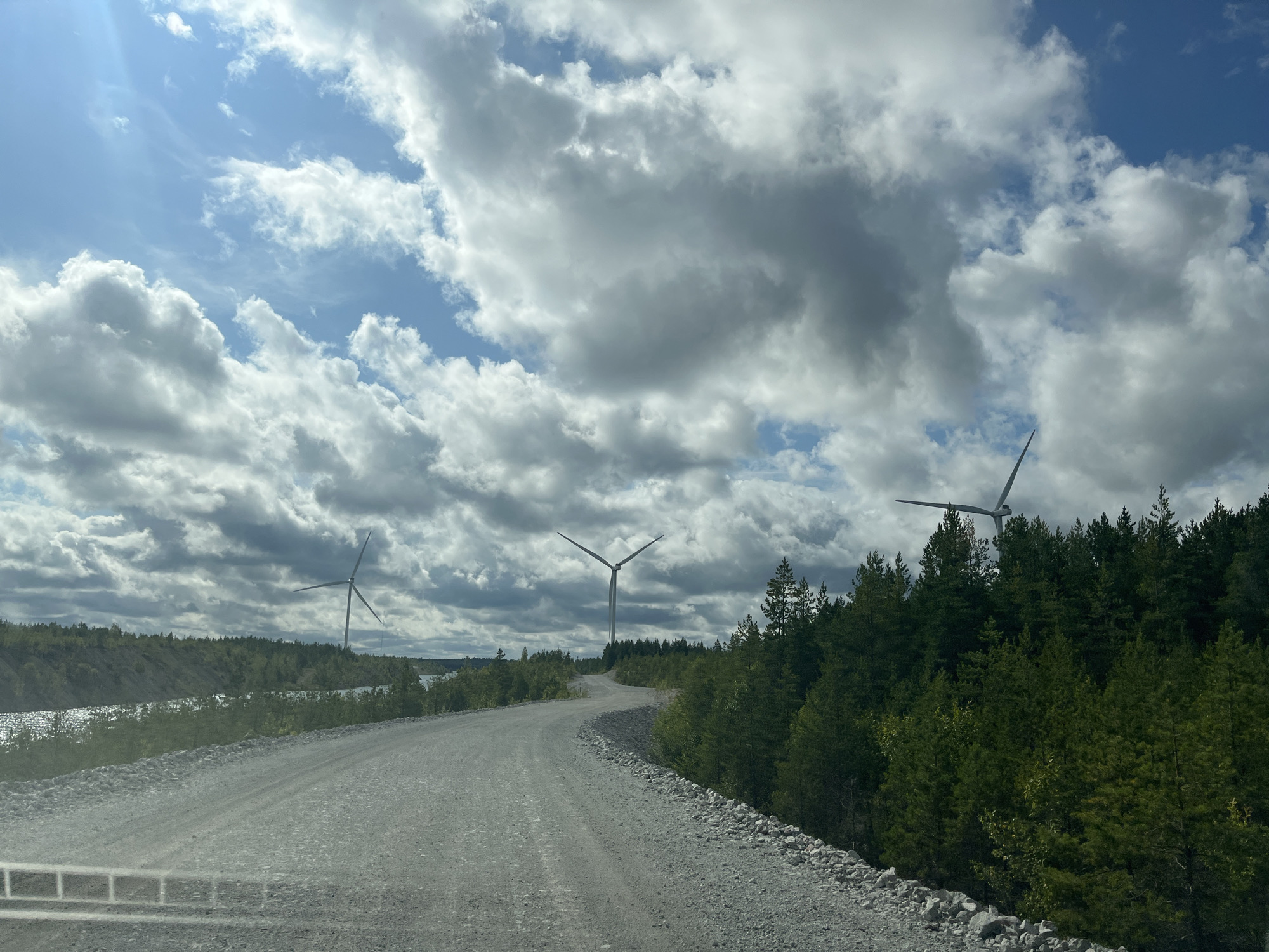 A winding gravel road leads through a sparsely populated landscape with wind turbines. The road is made of light-colored gravel and is bordered by trees on the right side of the image. In the distance, three wind turbines are visible, two of them on the left side of the road and one on the right. The sky is partly cloudy with patches of blue sky visible. A body of water is visible in the background on the left side of the image. The environment appears to be a rural or industrial area, possibly a former mining site given the barren ground on the left. The scene depicts a sunny day with scattered clouds, indicating a mild climate. The location is Aidu, Estonia.
