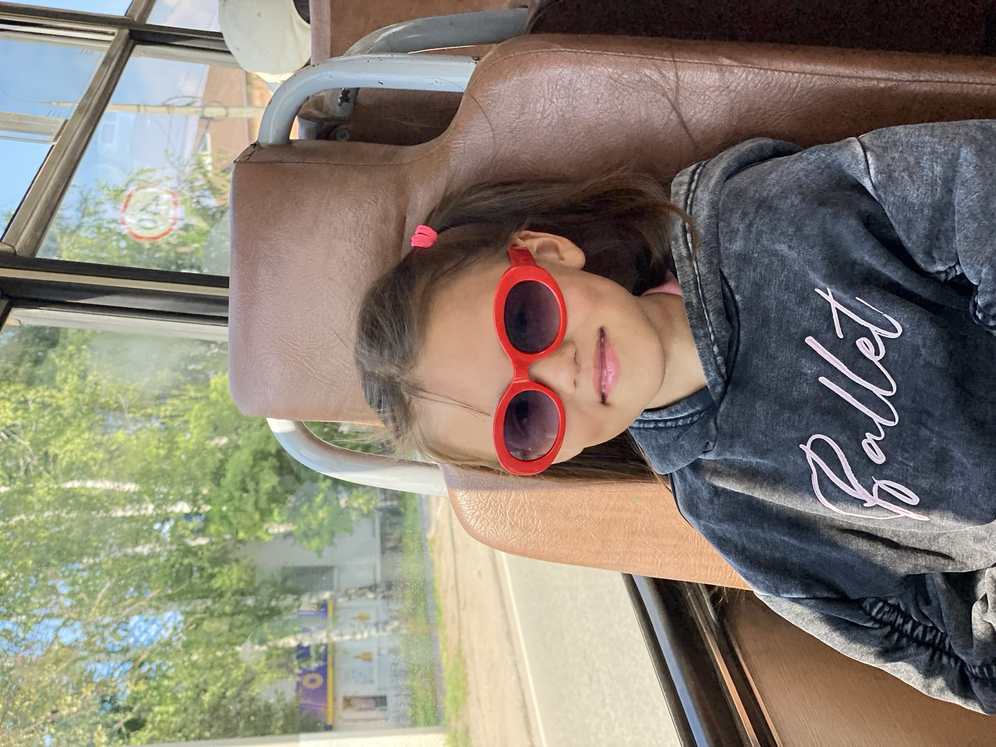 A young girl with brown hair tied into two pigtails and wearing bright red oval sunglasses is seated on a brown leather bus seat. She is wearing a dark gray hooded sweatshirt with the word "Ballet" printed in pink script on the front. The background through the bus window shows a sunny day with green trees and a blue sky, indicating an outdoor environment likely in a city like Chaikovsky, Russia. The bus interior is visible, with other seats and window frames present.