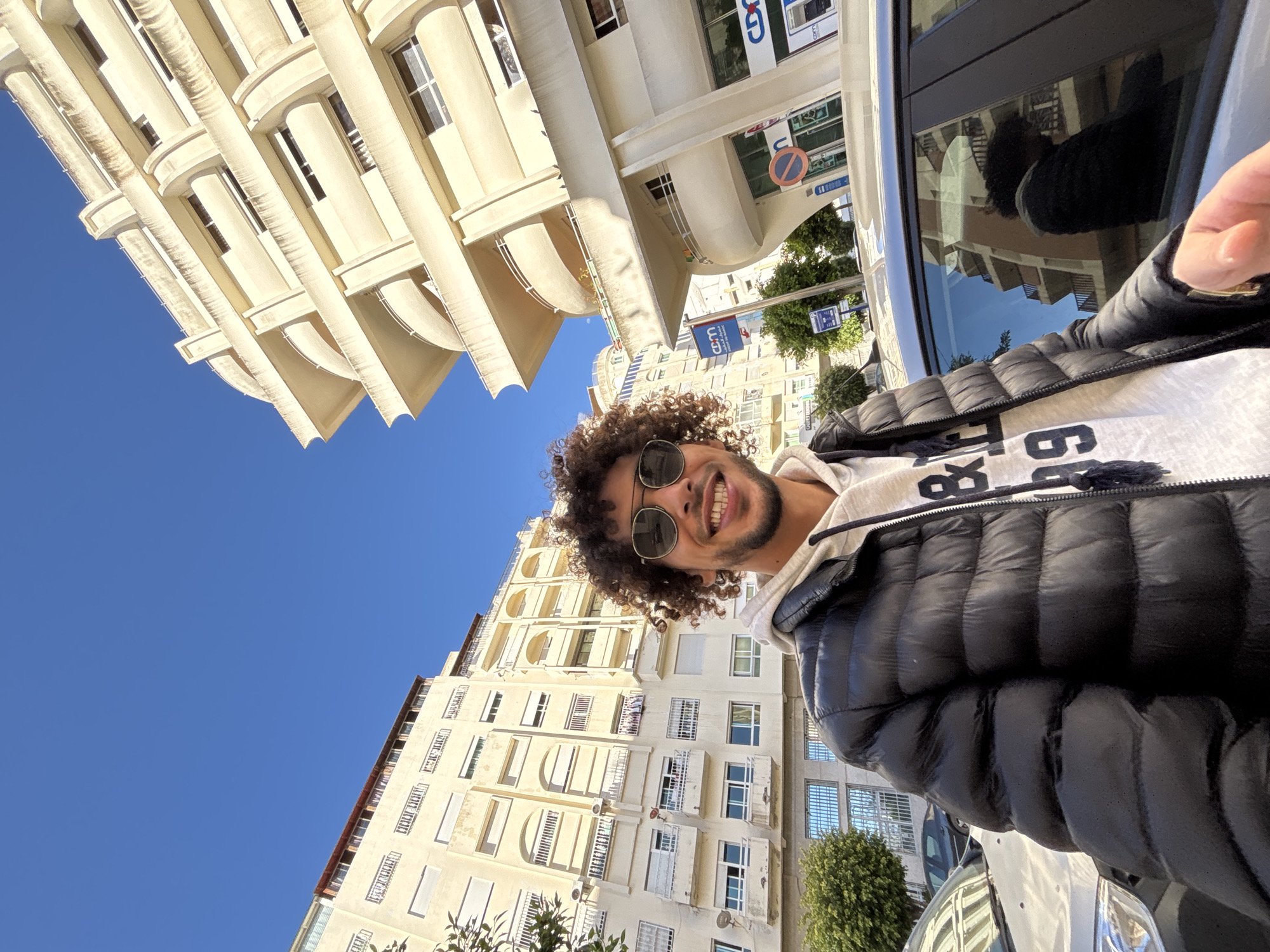A man with curly hair and round sunglasses stands next to a car, taking a selfie. He is wearing a black puffer jacket over a gray hooded sweatshirt. The background features buildings with cream-colored facades under a clear blue sky. The buildings have a mix of architectural styles with columns and balconies. Street signs in Arabic and French are visible. The location appears to be an urban street in the city of Tanger طنجة, Morocco.