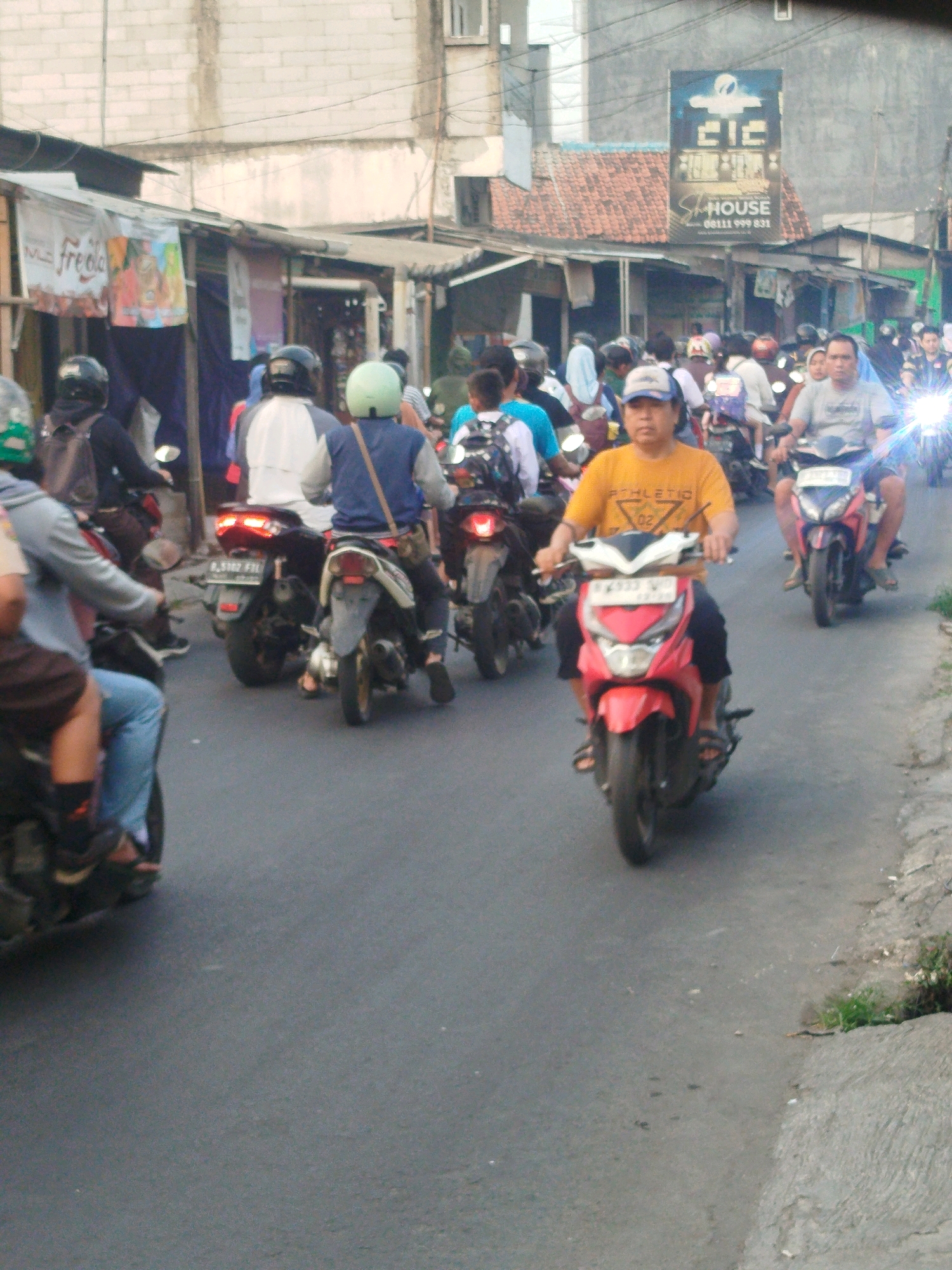 The image depicts a street scene in Karangsatria, Indonesia, characterized by a moderate amount of traffic, primarily consisting of motorcycles. The architecture visible includes buildings with visible brickwork and some shops with awnings. Several individuals are on motorcycles, either as drivers or passengers, navigating the paved road. One individual in the foreground is wearing a yellow t-shirt and riding a red scooter. The overall atmosphere suggests a typical day in an urban or semi-urban Indonesian setting, with a focus on motorcycle transportation.
