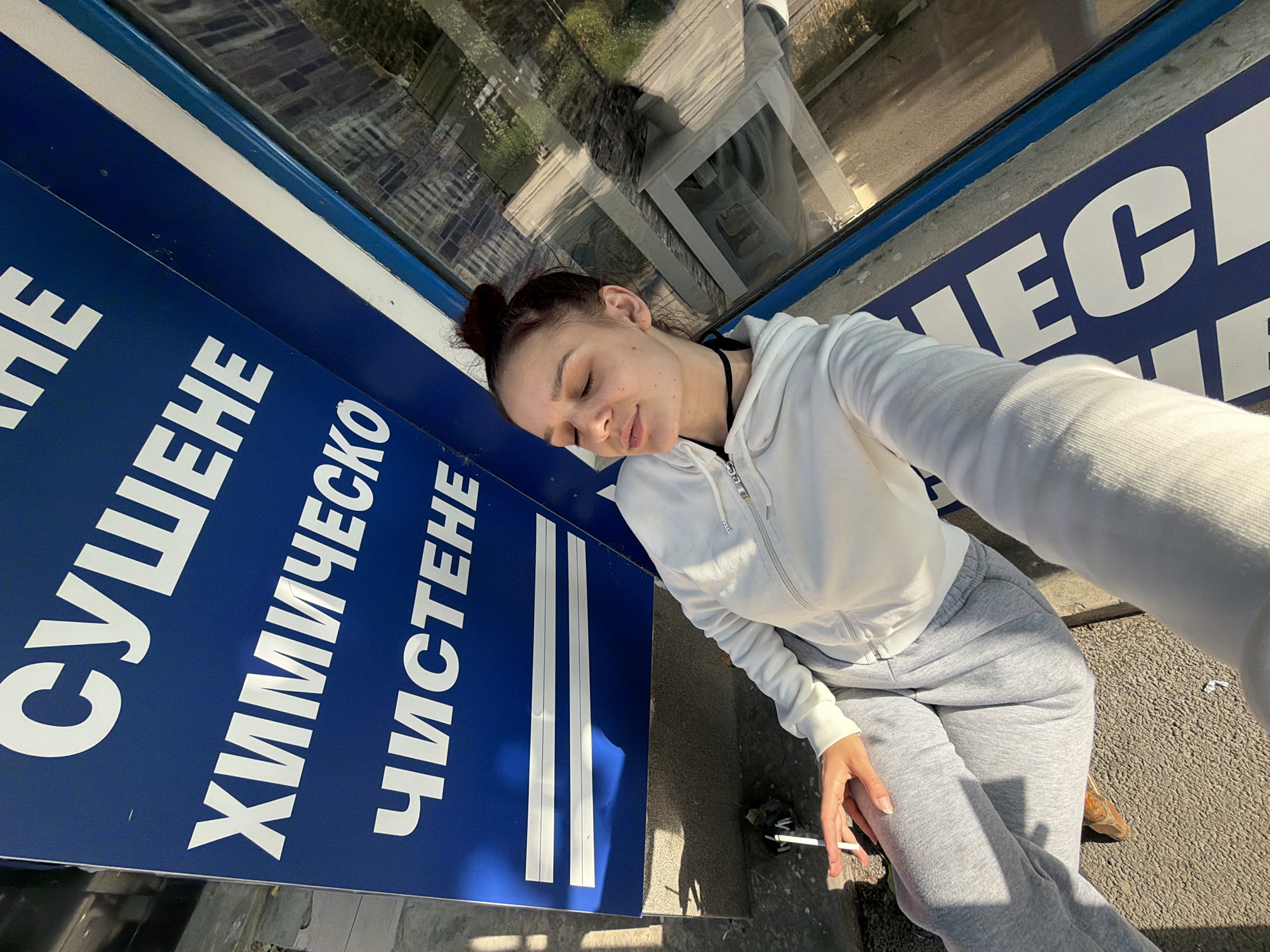 A woman with her eyes closed is sitting outdoors, holding a cigarette. She is wearing a white hooded sweatshirt and grey sweatpants. The background features a blue sign with white Cyrillic lettering. The sign appears to be for a dry cleaning service, with the words "СУШЕНЕ" (Drying), "ХИМИЧЕСКО" (Chemical), and "ЧИСТЕНЕ" (Cleaning) visible. Sunlight casts shadows on the ground. A reflection in a glass surface behind the woman shows buildings and greenery. The scene is suggestive of a public space in Sofia, Bulgaria, given the use of Cyrillic and the typical urban setting.