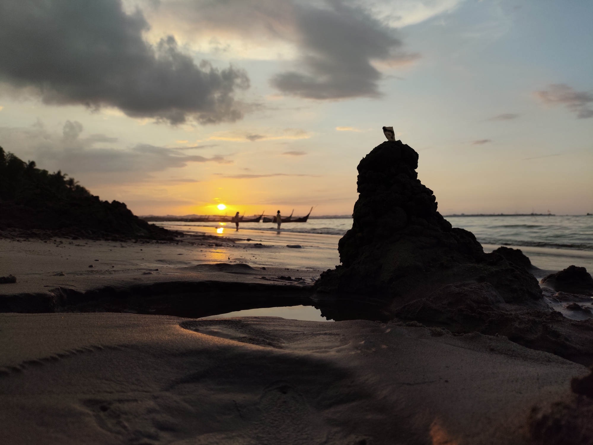 The image captures a silhouetted beach scene at sunset, likely in Lhokseumawe, Indonesia, given the presence of traditional outrigger boats in the water. The foreground features a sandy beach with a small pool of water reflecting the sky. To the right, a dark, jagged rock formation stands prominently, topped with a small, light-colored object that appears to be a piece of paper or plastic. In the middle ground, the setting sun casts a warm glow across the calm sea. Several outrigger boats, each with figures standing in them, are visible on the water, suggesting fishing or travel activities. In the background, the horizon is marked by the sea meeting the sky, with a faint line of what could be distant structures or land. The sky itself is a blend of soft oranges, yellows, and blues, with scattered clouds in varying shades of grey and white, indicating the transition from day to night. The overall atmosphere is serene and picturesque, typical of a tropical coastal sunset.