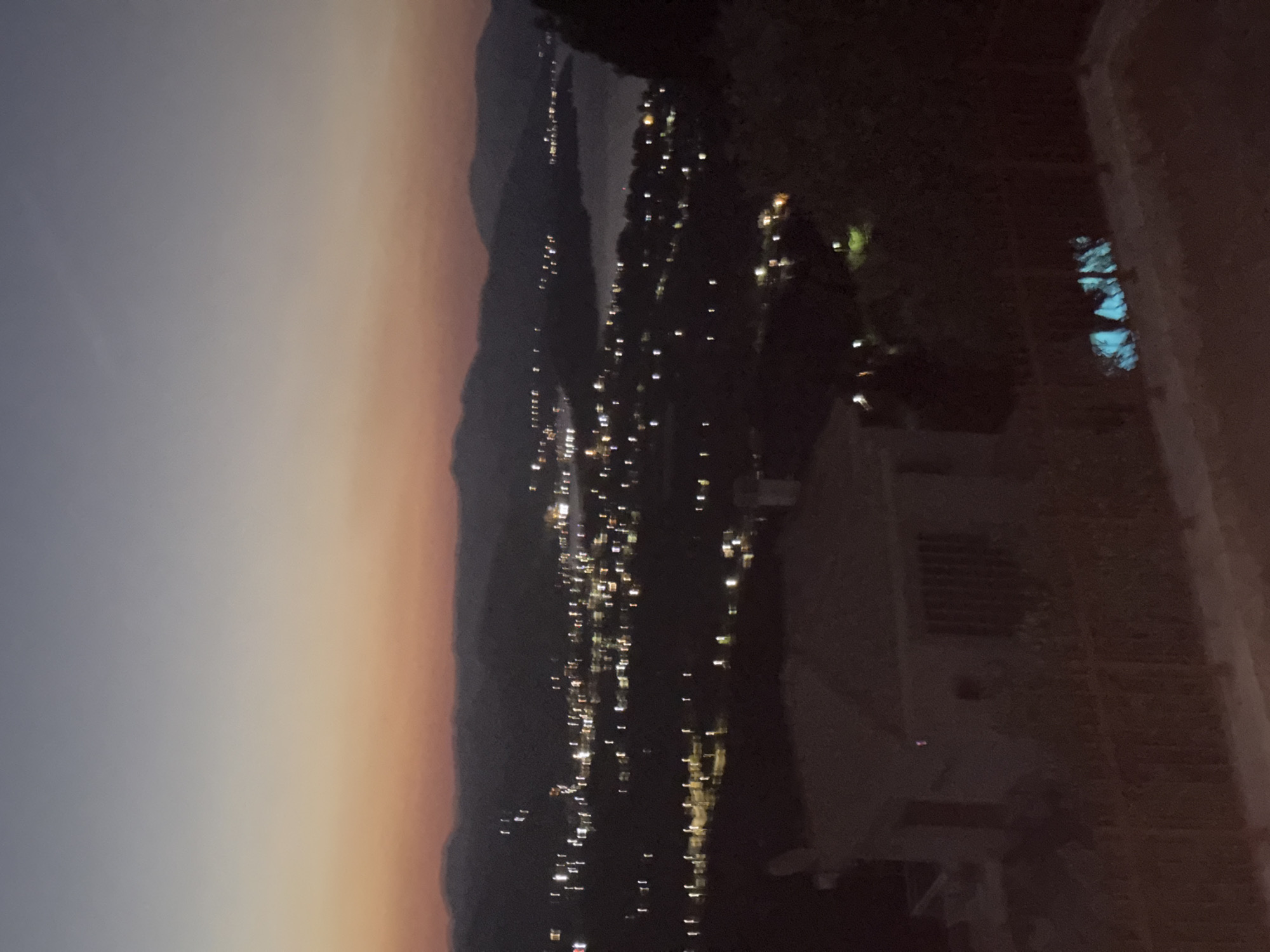 The image captures a twilight view of the Municipal Unit of Asini, Greece, with a town illuminated by lights in the distance. The sky transitions from a pale blue at the top to hues of orange and pink near the horizon, suggesting the sun has recently set. Hills or mountains form a dark silhouette against the sky. In the foreground, a low-lying structure, possibly a house or small building, is visible. Further out, the lights of the town and what appears to be a body of water are discernible. There is a fenced area in the foreground, and in the lower right corner, a brightly lit section of what could be a swimming pool or water feature. The overall scene is dimly lit, indicative of dusk or early evening.