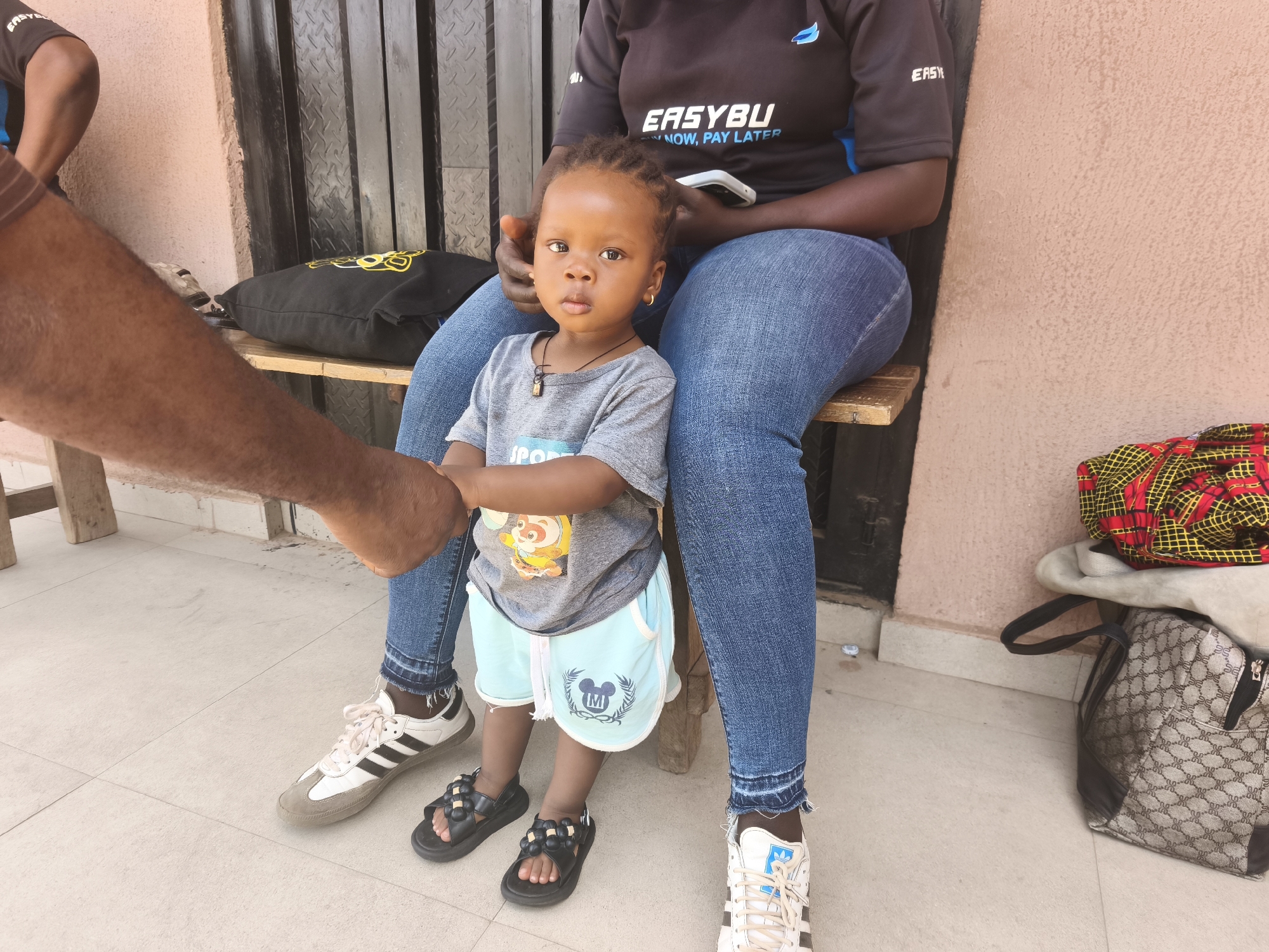 A young child stands in front of an adult seated on a bench in Jos, Nigeria. The child, wearing a gray t-shirt, blue shorts, and black sandals, is holding the hand of another adult whose arm is visible on the left. The seated adult is wearing jeans, a dark t-shirt with "EASYBUY" printed on it, and white sneakers. A black pillow is on the bench behind the adult. The background includes a textured wall and a patterned bag on the ground. The floor is tiled.
